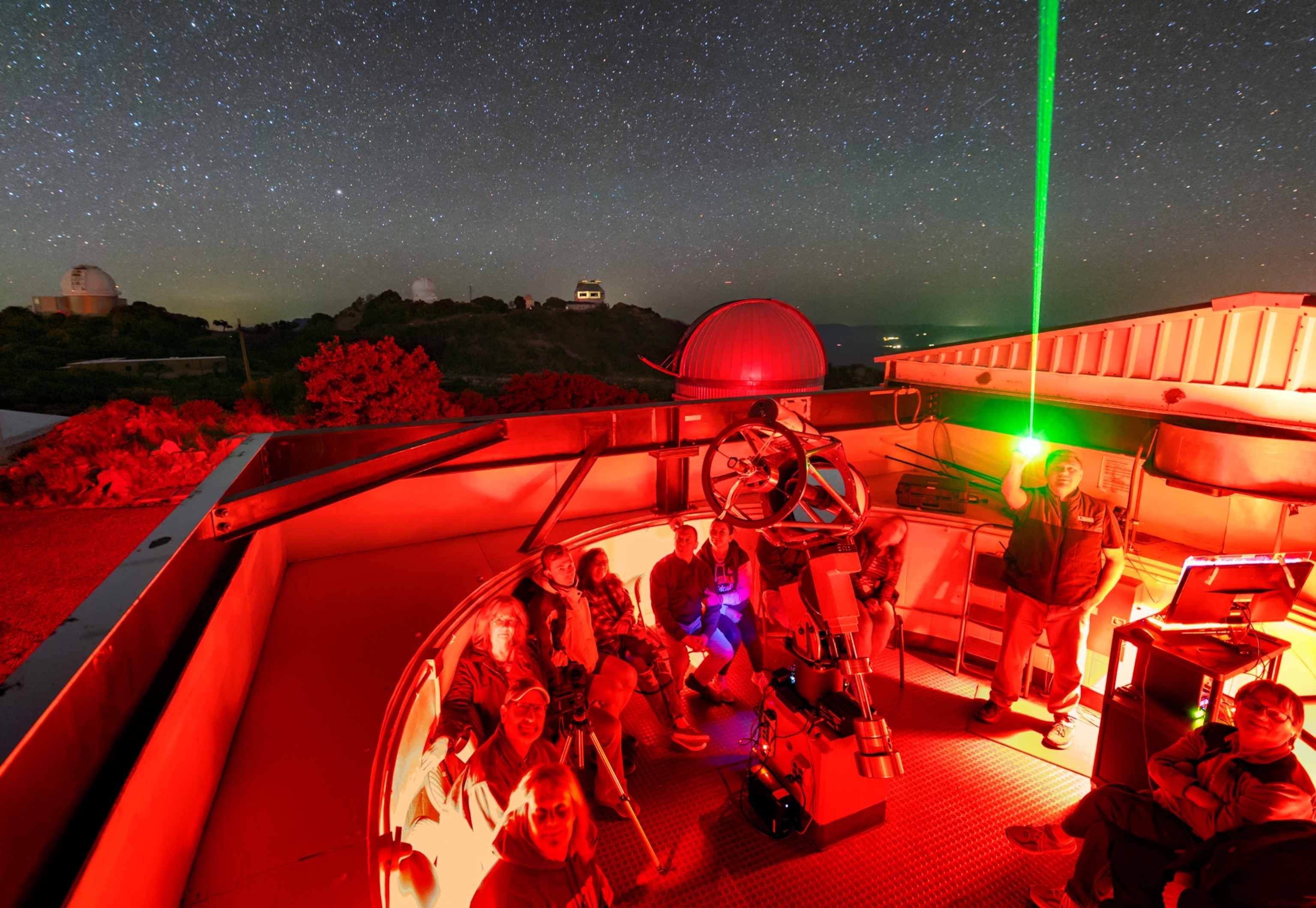 A group of people inside an observatory, lit by red lights, gaze upwards. A green laser points at the starry night sky