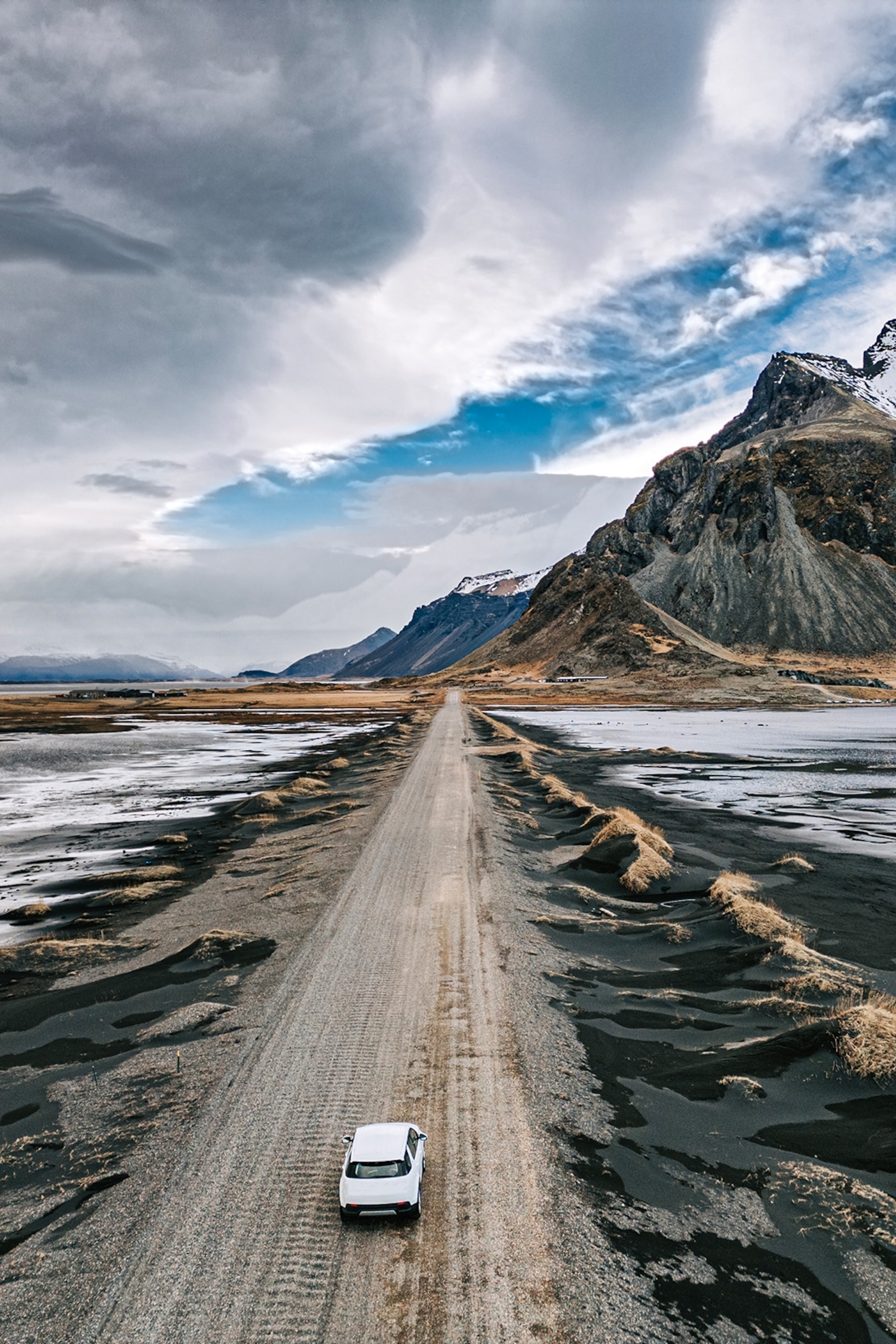 A single car making its way down a muddy road splitting a black sand beach, snow-capped and rugged mountains rise up in the distance.