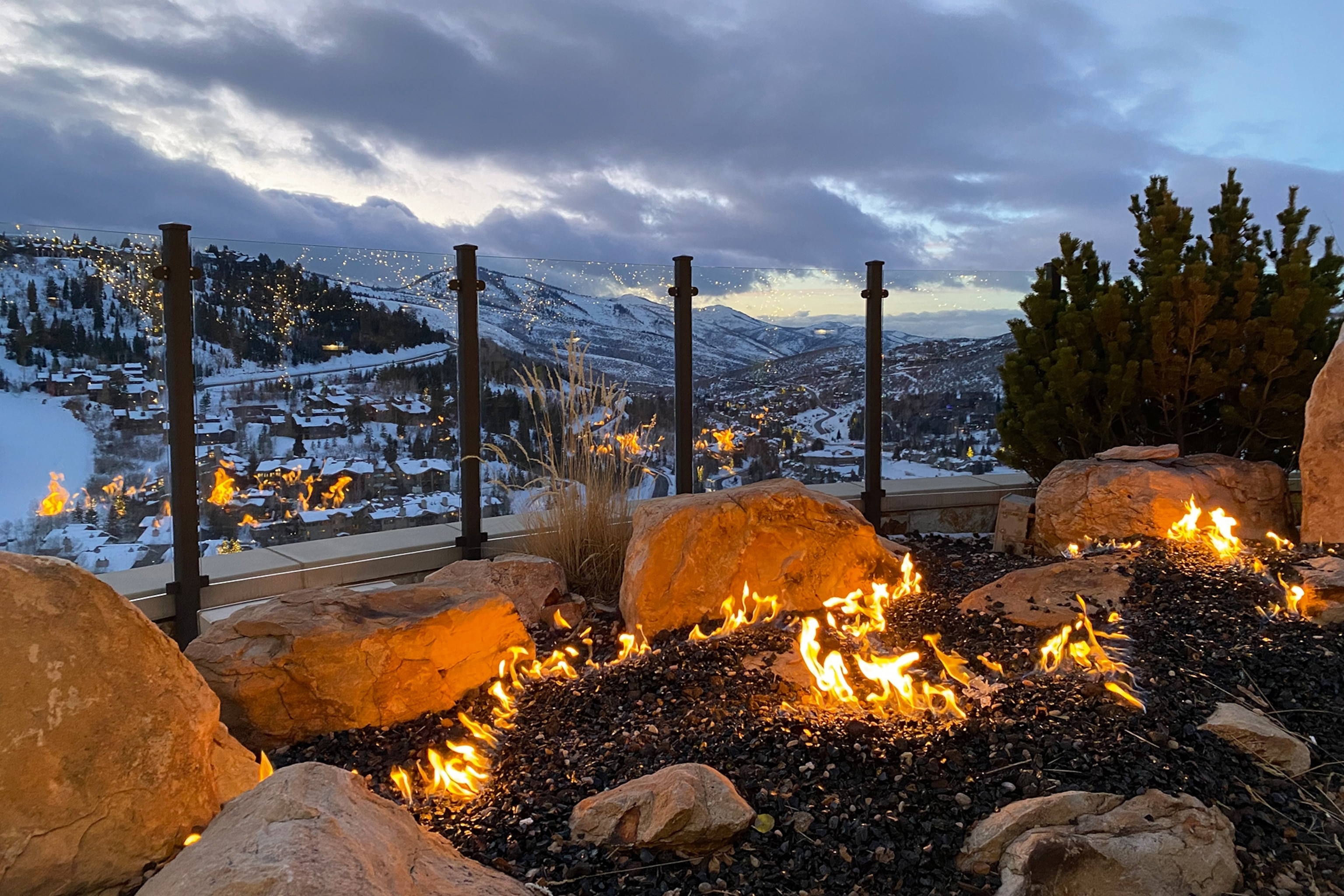 A nighttime view over a snowy mountain valley dotted with illuminated houses and a lit firepit in the foreground.