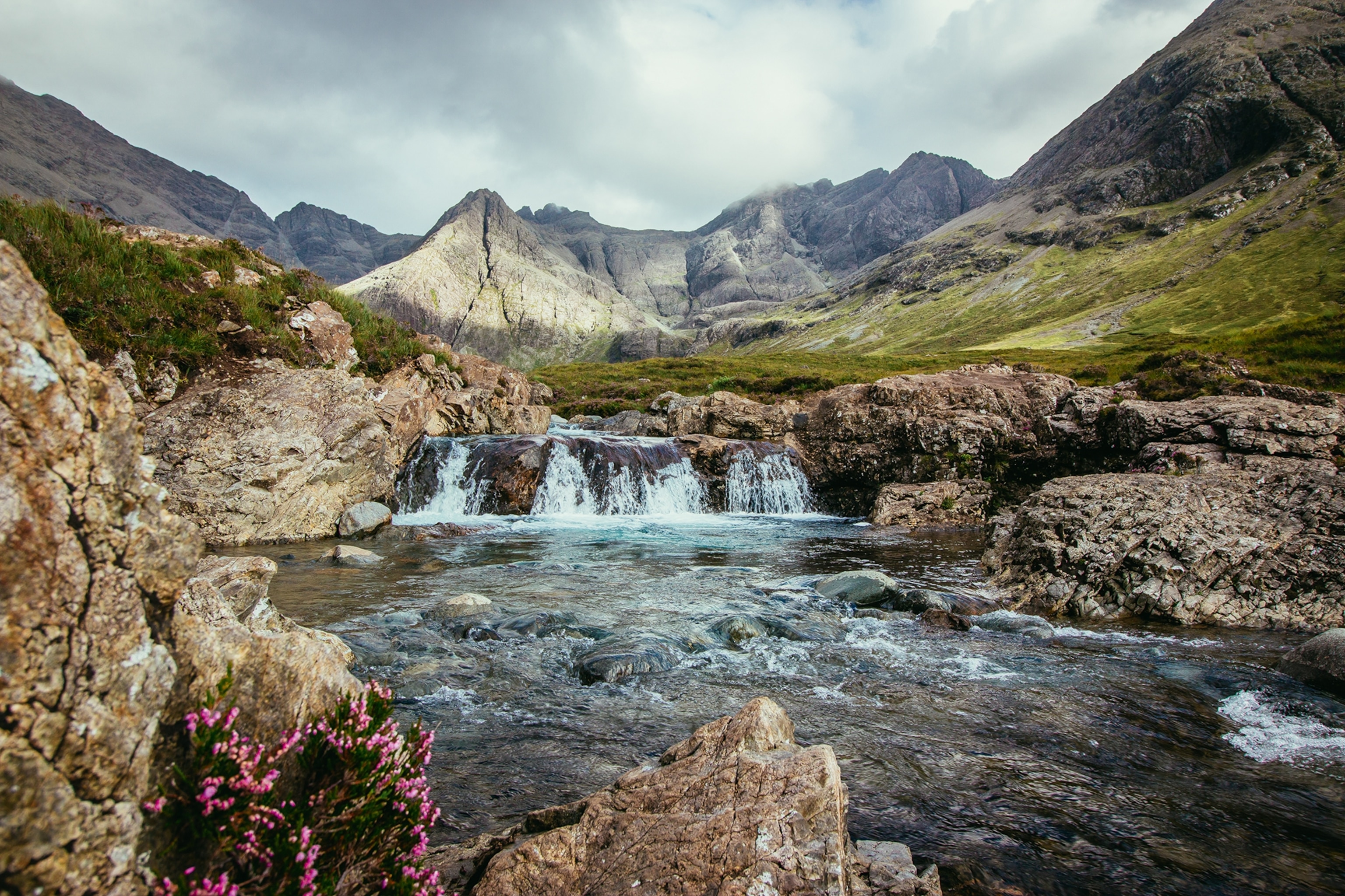 A wild and rocky pool fed by a multi-tiered yet low waterfall with an impressive mountain range in the background.