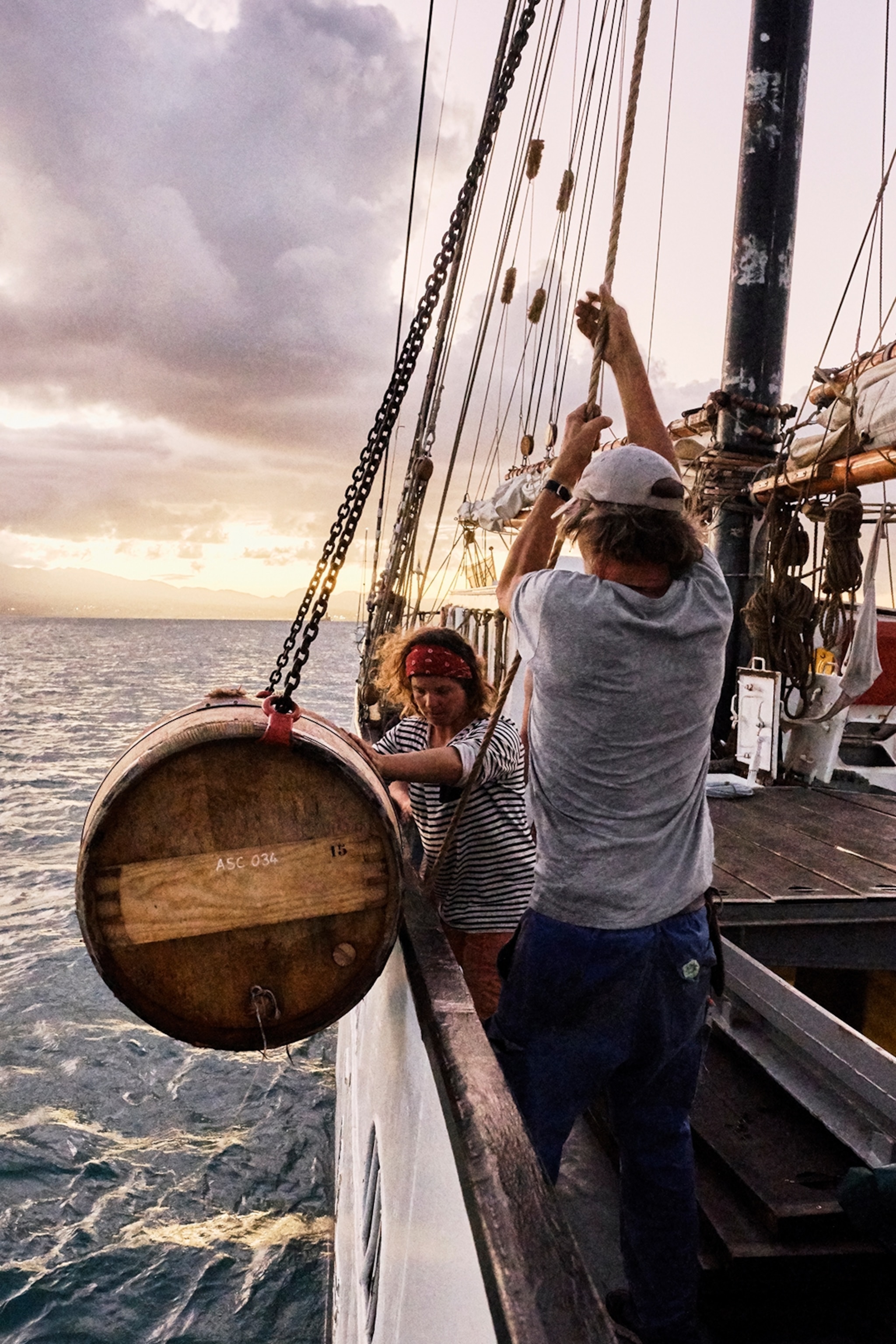 A crew o lowering barrels filled with rum from sailing boat down into the ocean below with ropes.