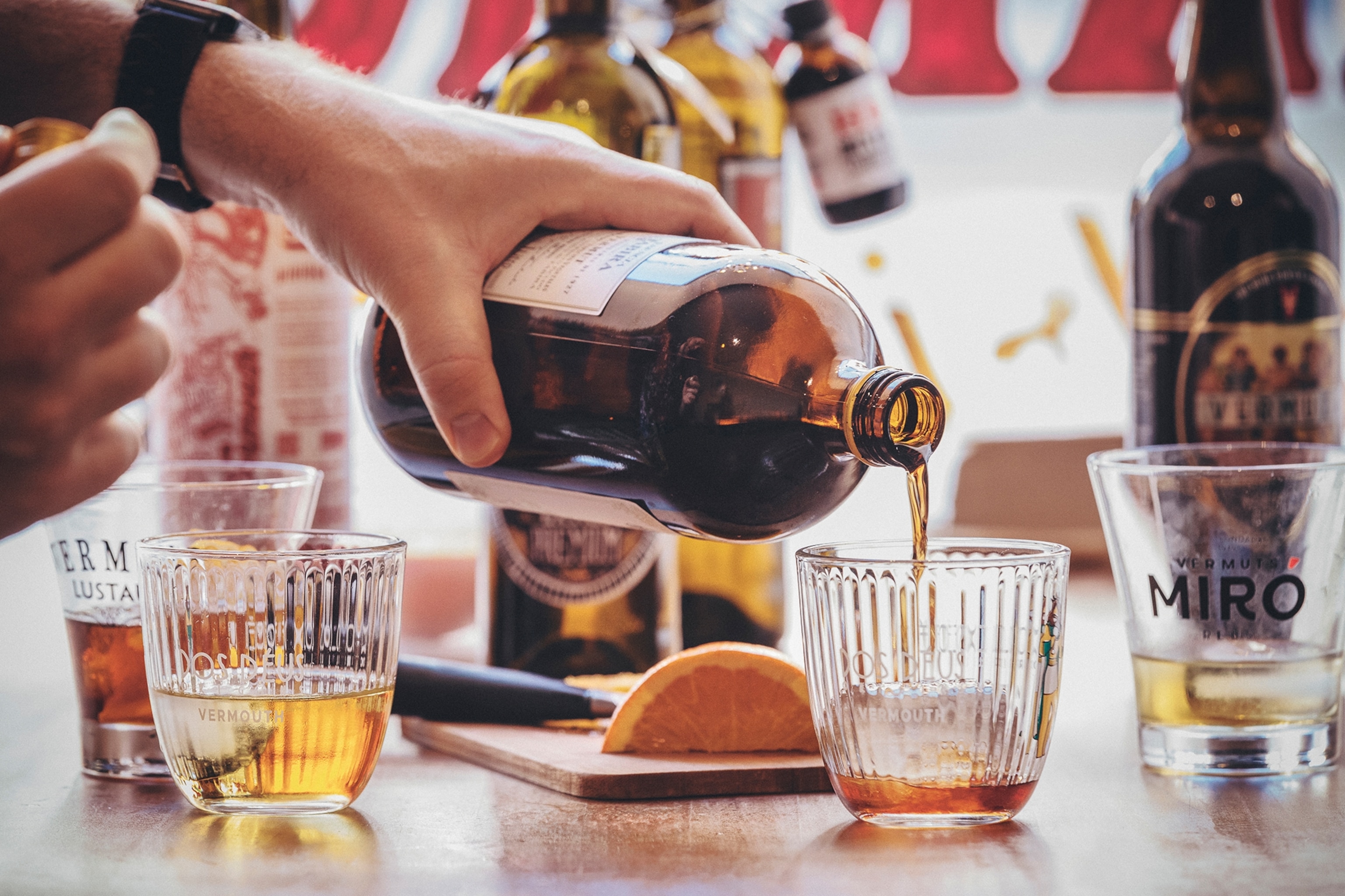 A close-up of a person pouring a shot of vermouth on a table with other glasses and bottles.