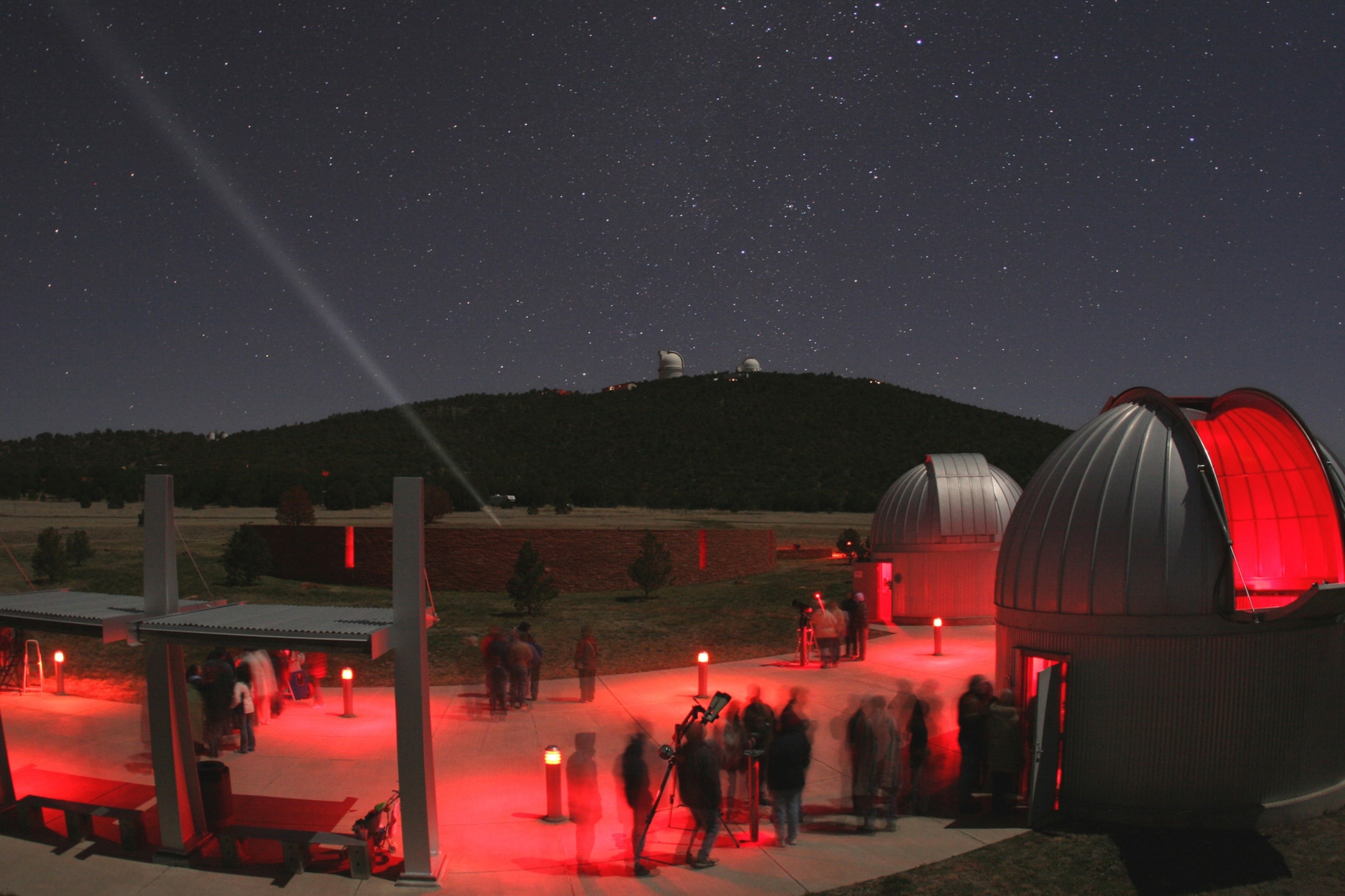 People walking around an observatory outside under a dark sky filled with stars. The ground is lit up with red lights.