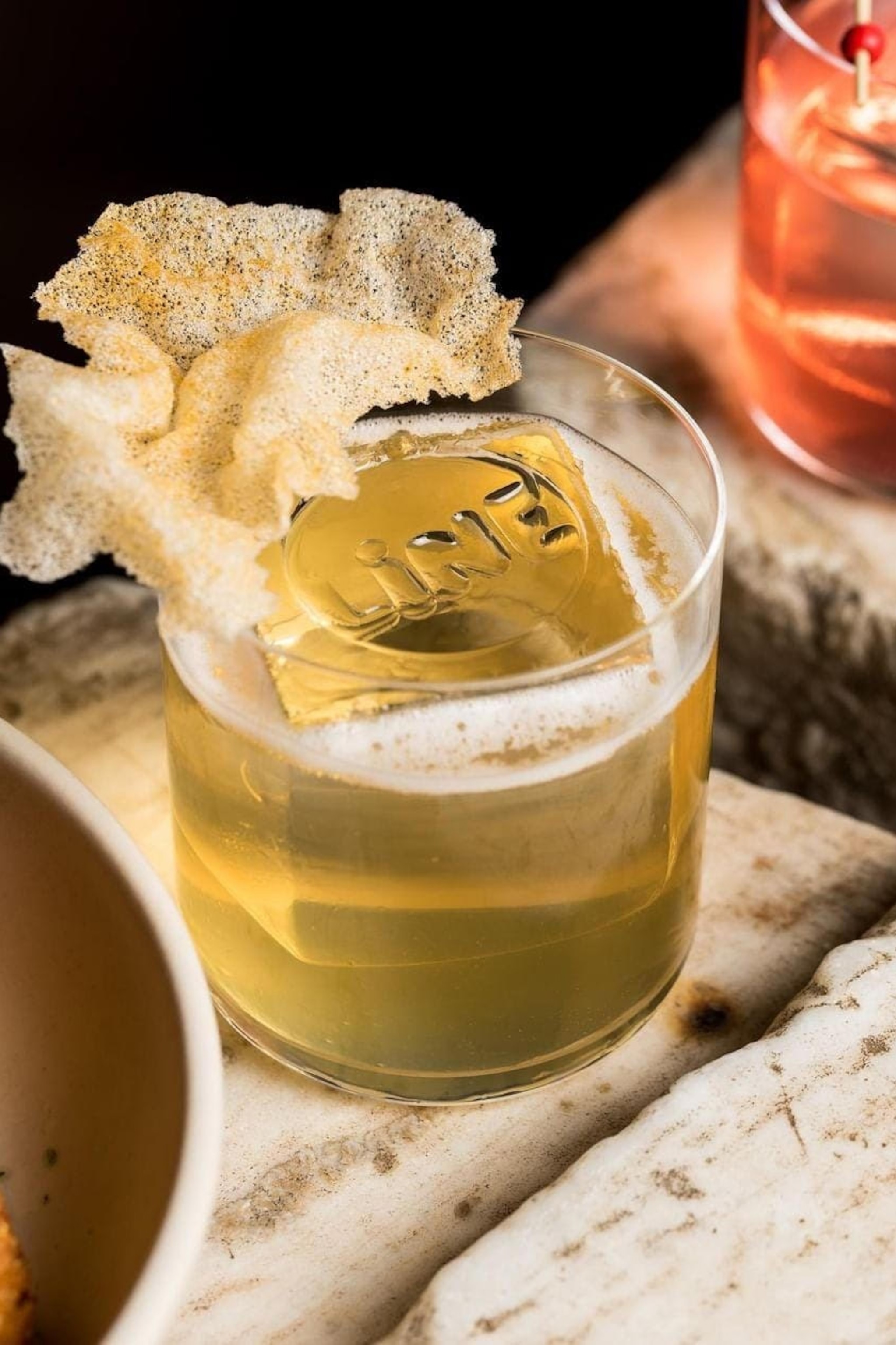 A close-up of a cocktail in a tumbler with a square ice cube and a poppadum crisp balance on the glass rim.