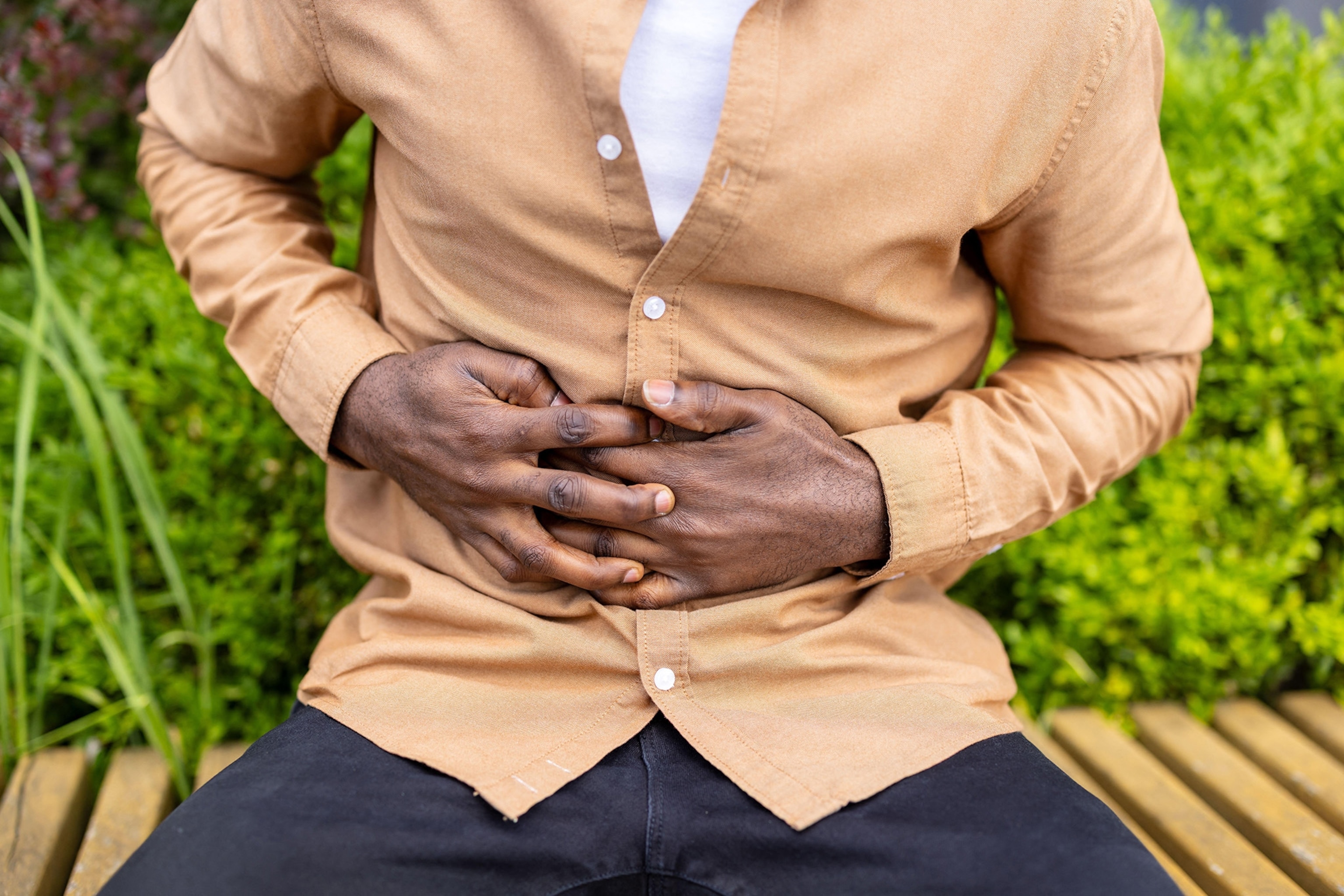 A man wearing a tan button down with a white t-shirt clutches his abdomen with both hands while sitting down on a bench outside.