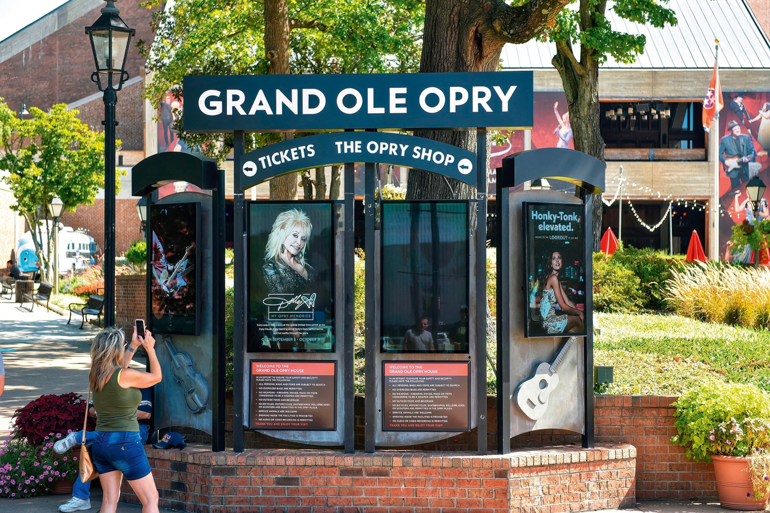 A female tourist taking a picture of a four-fold sign on a street corner, advertising tickets to a radio show.