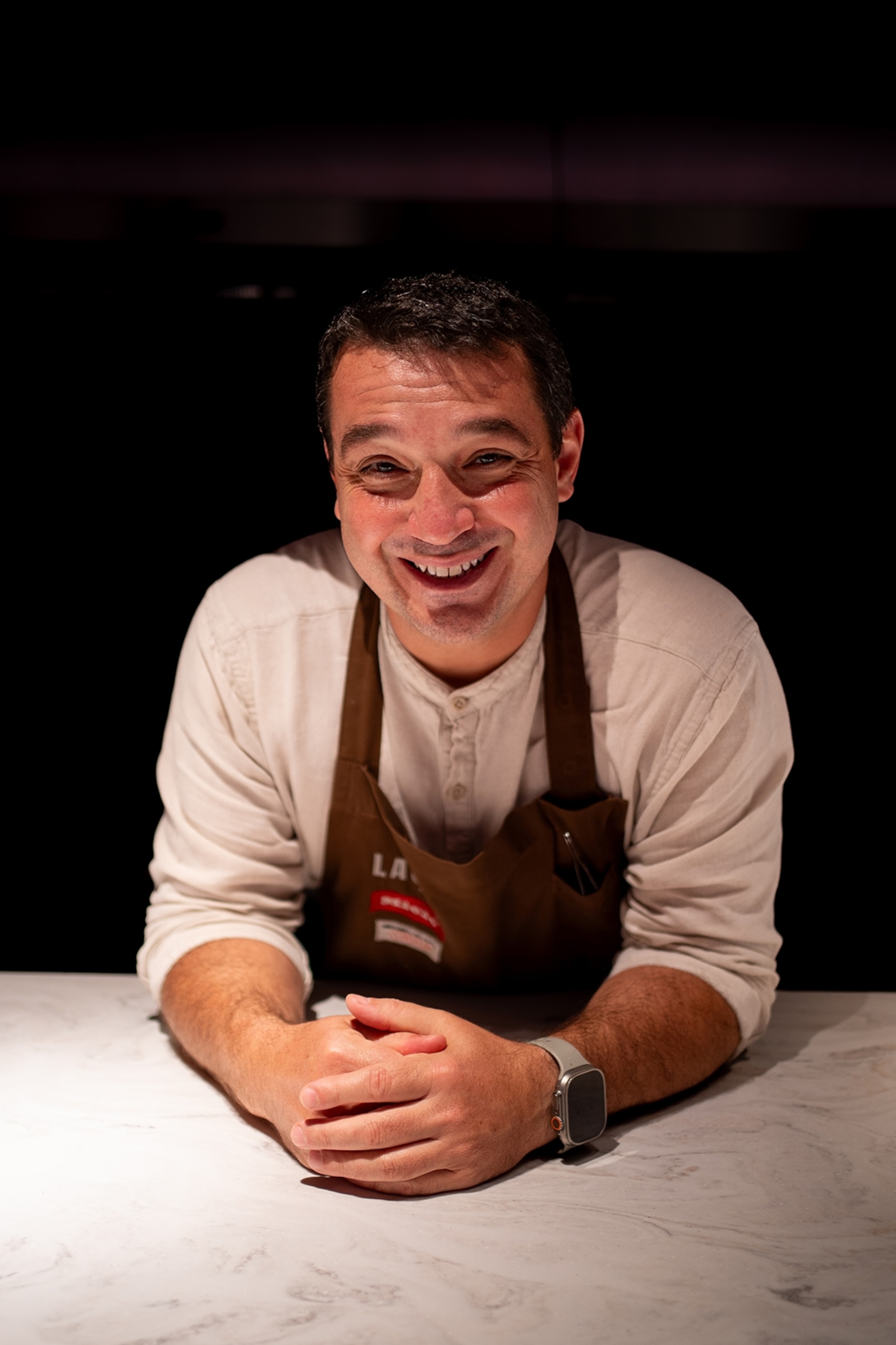 A young, male chef leaning forward onto a kitchen counter and smiling for the camera.