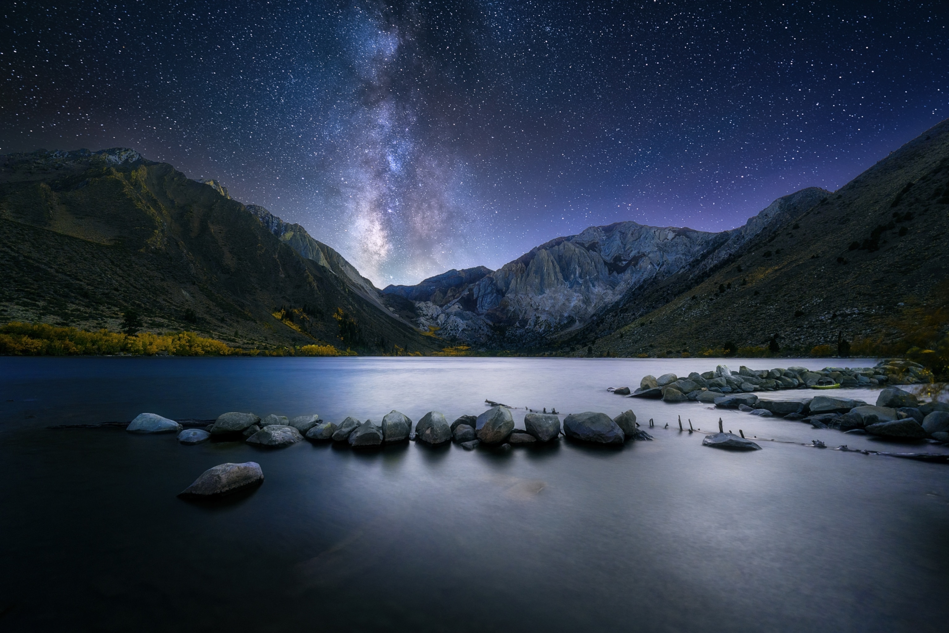 A calm mountain lake reflects the night sky, with rocky shorelines in the foreground and the Milky Way shining above rugged peaks in the distance.