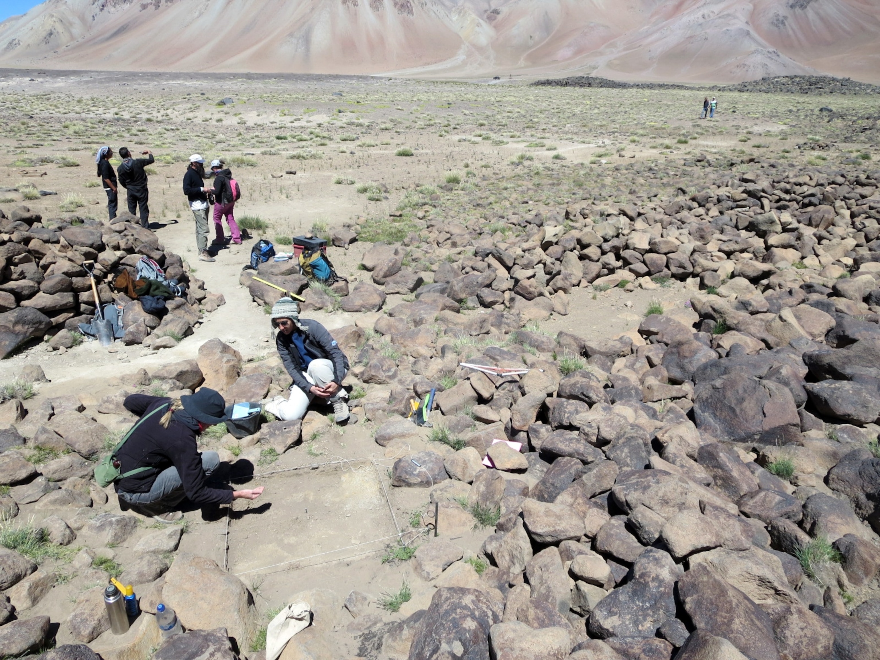 Archaeologists work at a dig site surrounded by rocks in a mountainous desert landscape. A group stands in the background, with dusty terrain all around.