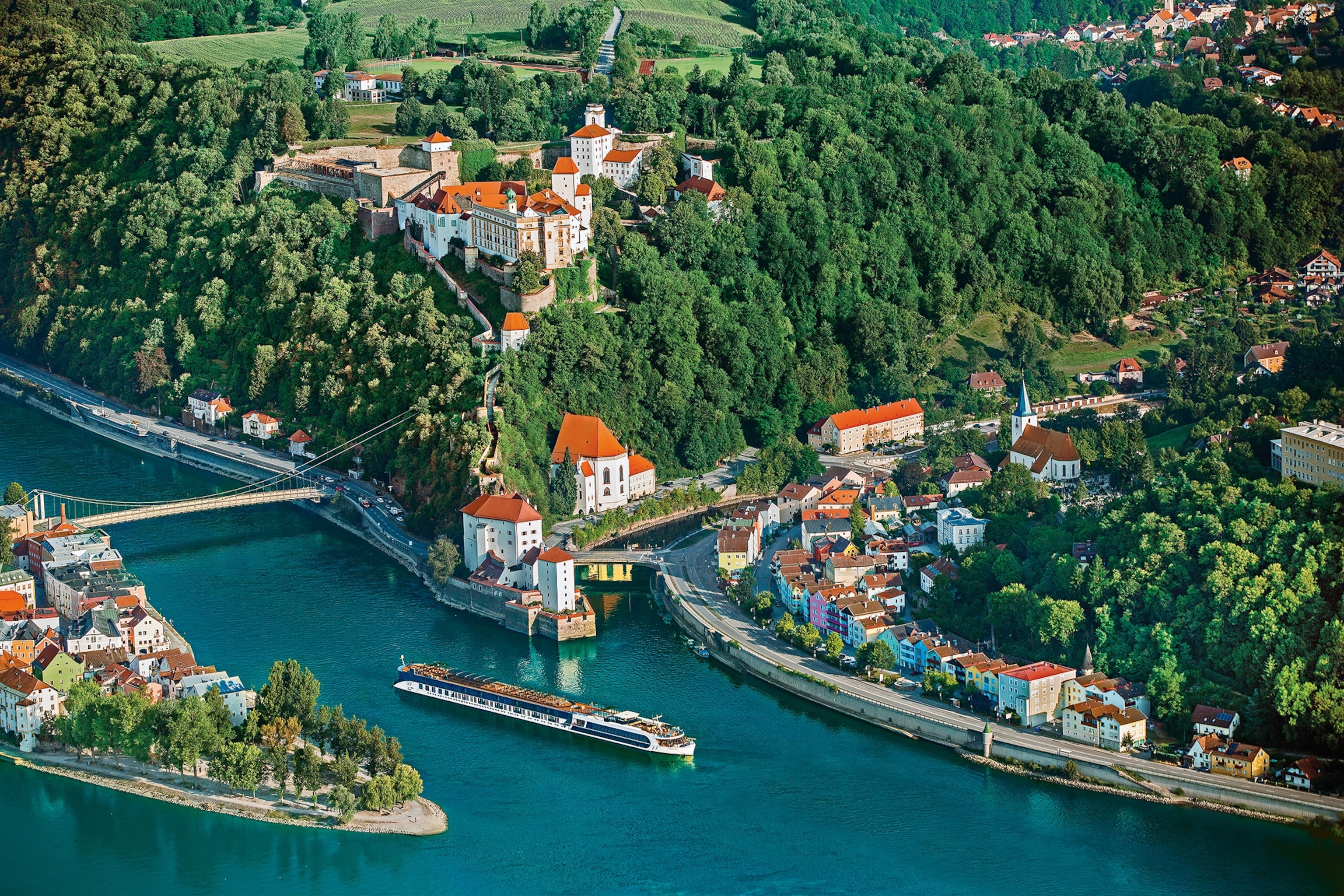 An aerial shot of a forking river in Germany with baroque-style houses and a small castle at the top of a forested hill.