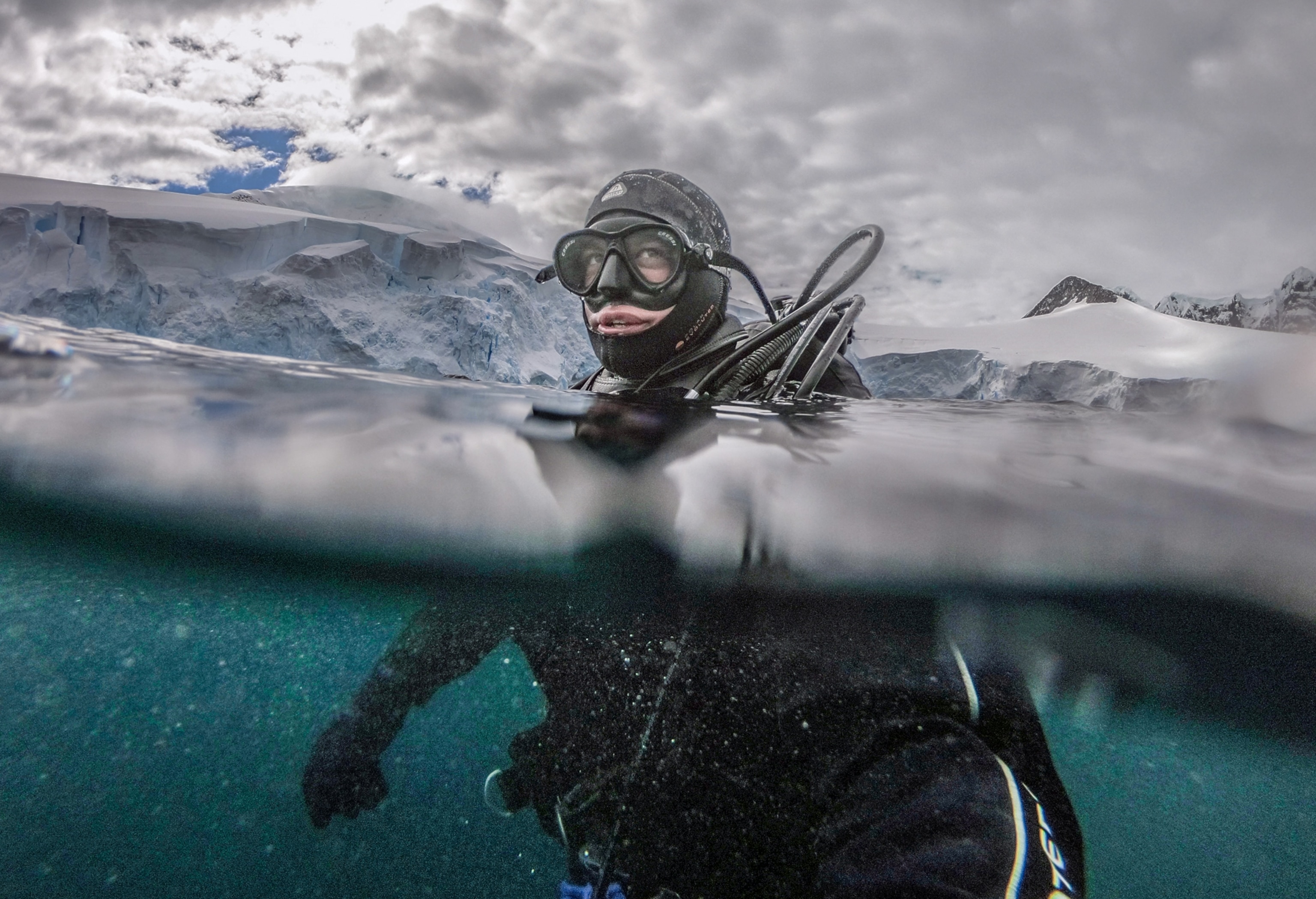 Man in a drysuit and mask floating upright with his had above and body under crystal clear water.