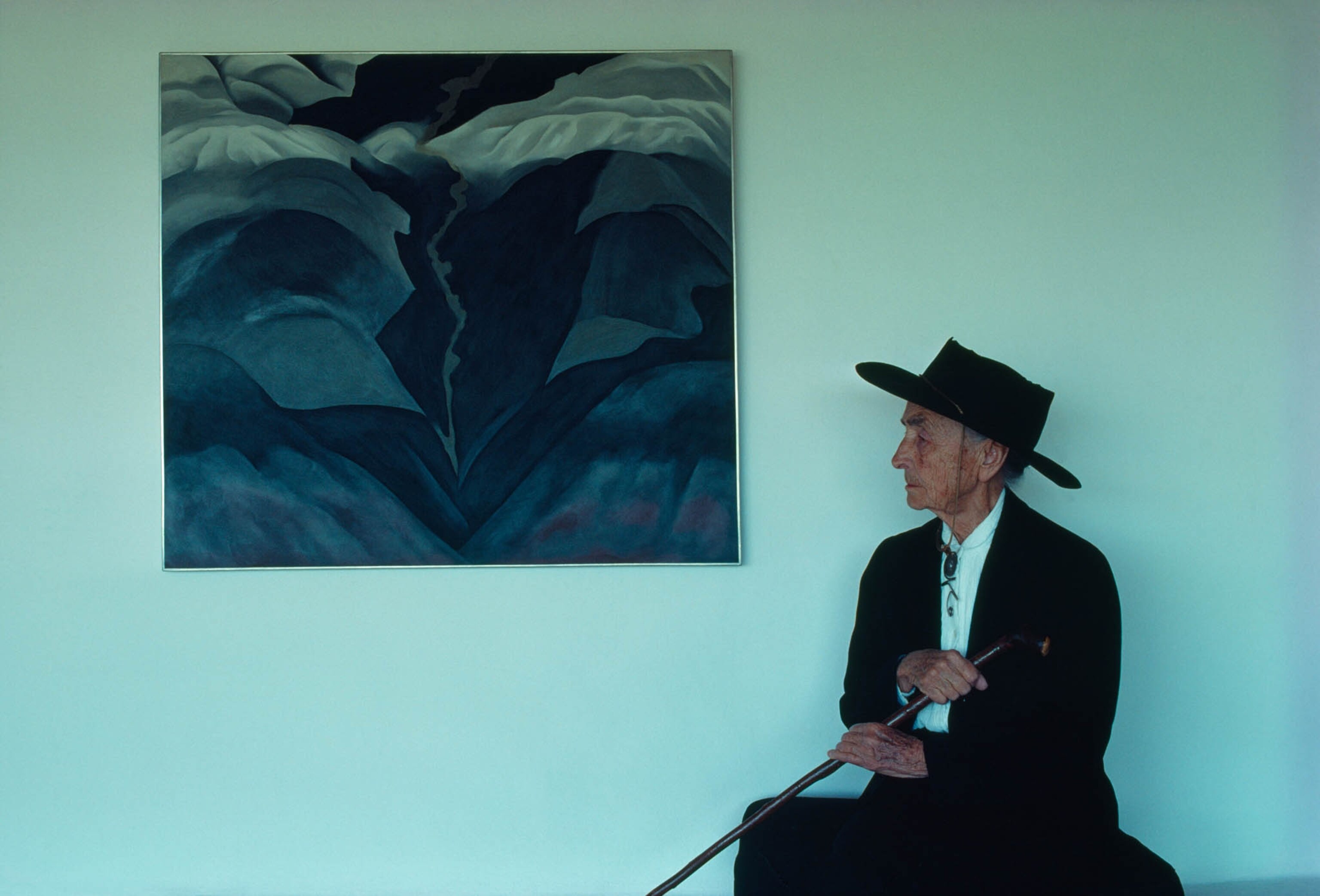 Artist Georgia O'Keeffe wearing a wide-brimmed black hat and sitting next to her painting titled Black Place III.