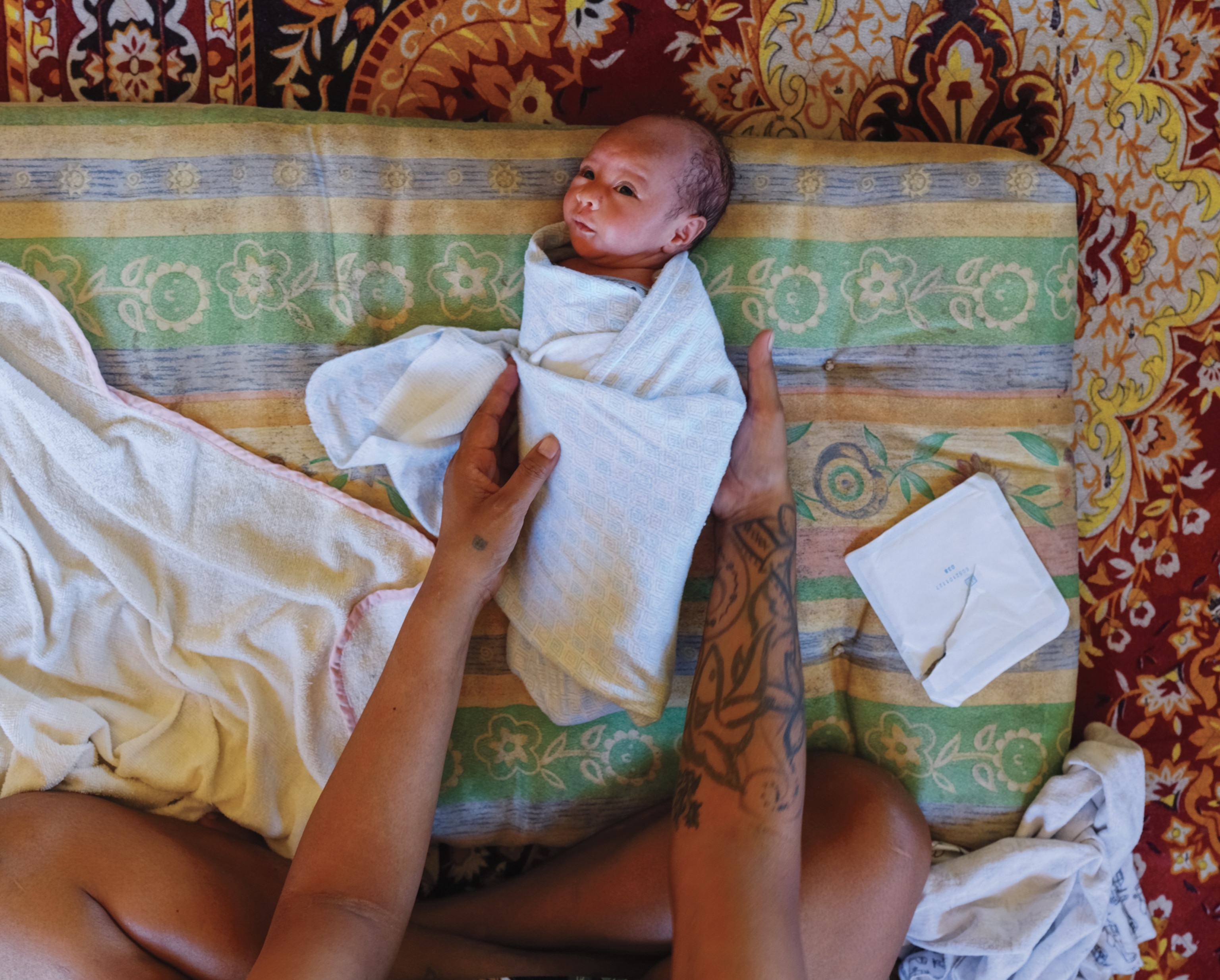 A baby lays on a pad on the ground as he gets swaddled in a white blanket.