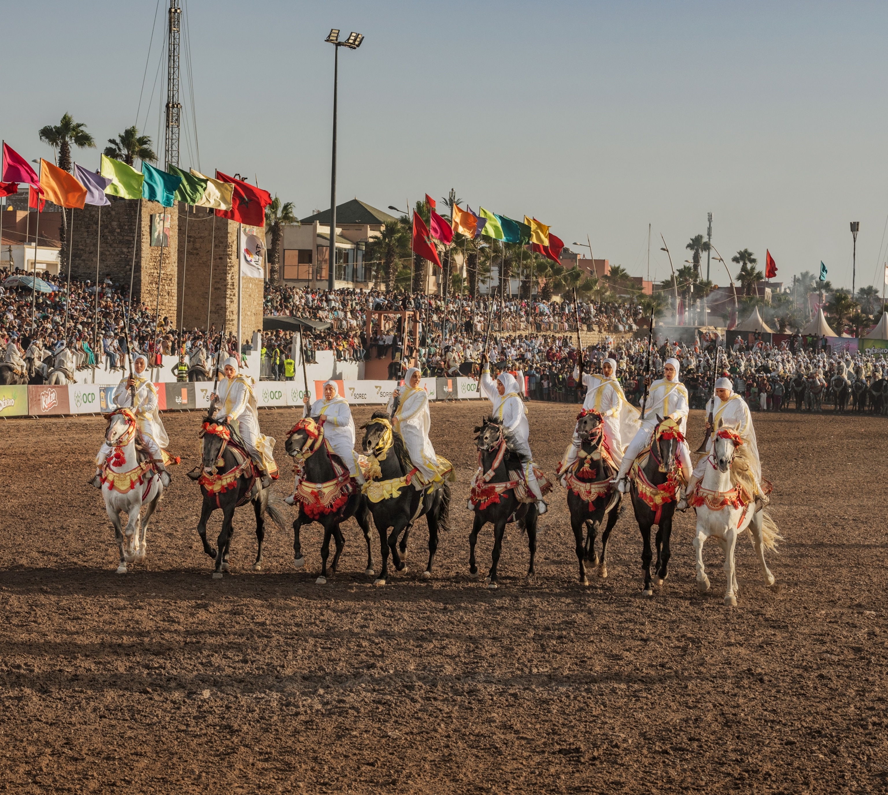 A group of 8 riders on horses move in a horizontal line towards the camera on a dirt arena as people observe in the crowd around and behind them.