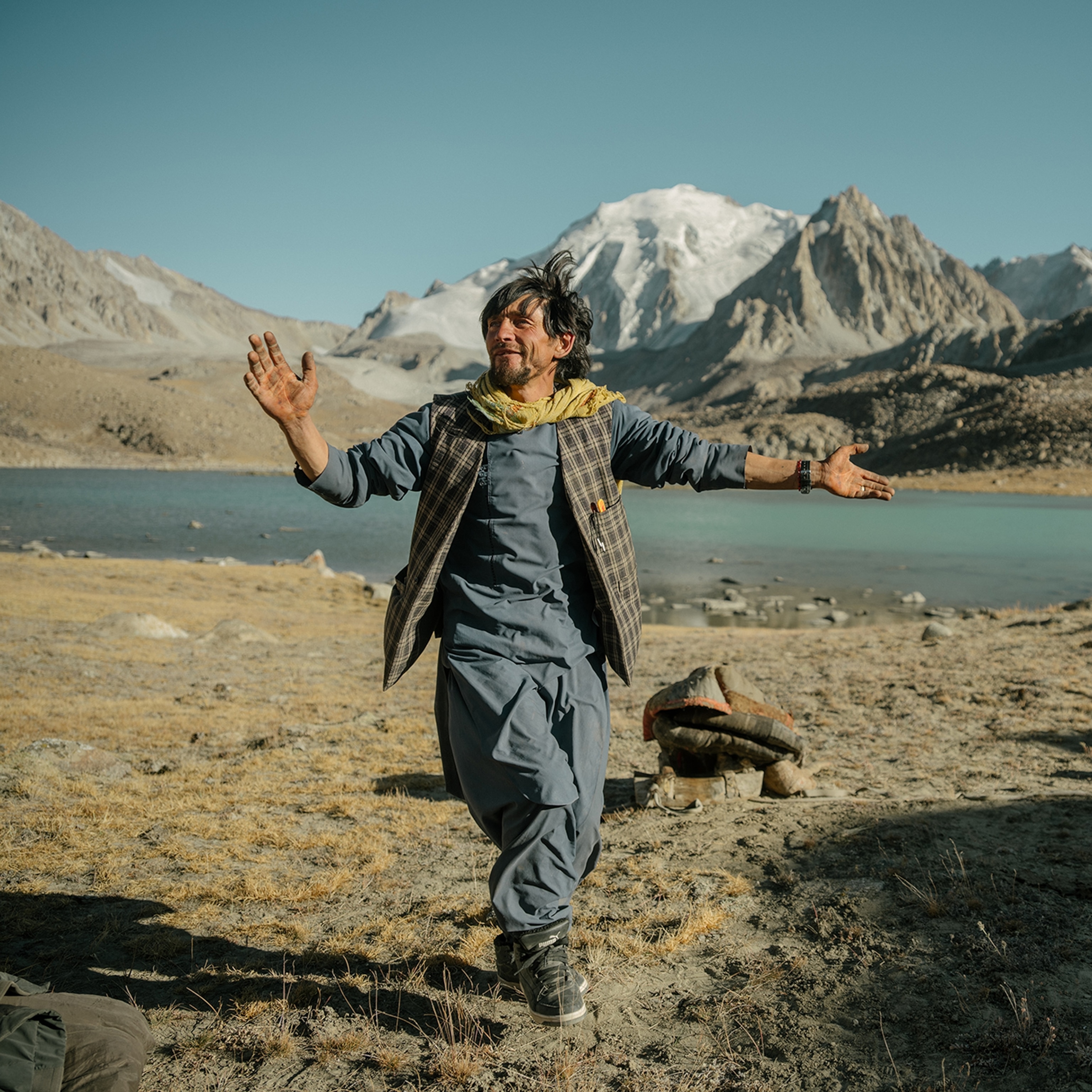 A man wearing blue clothes and a yellow scarf dances in a field with a lake and mountains in the distance behind him.