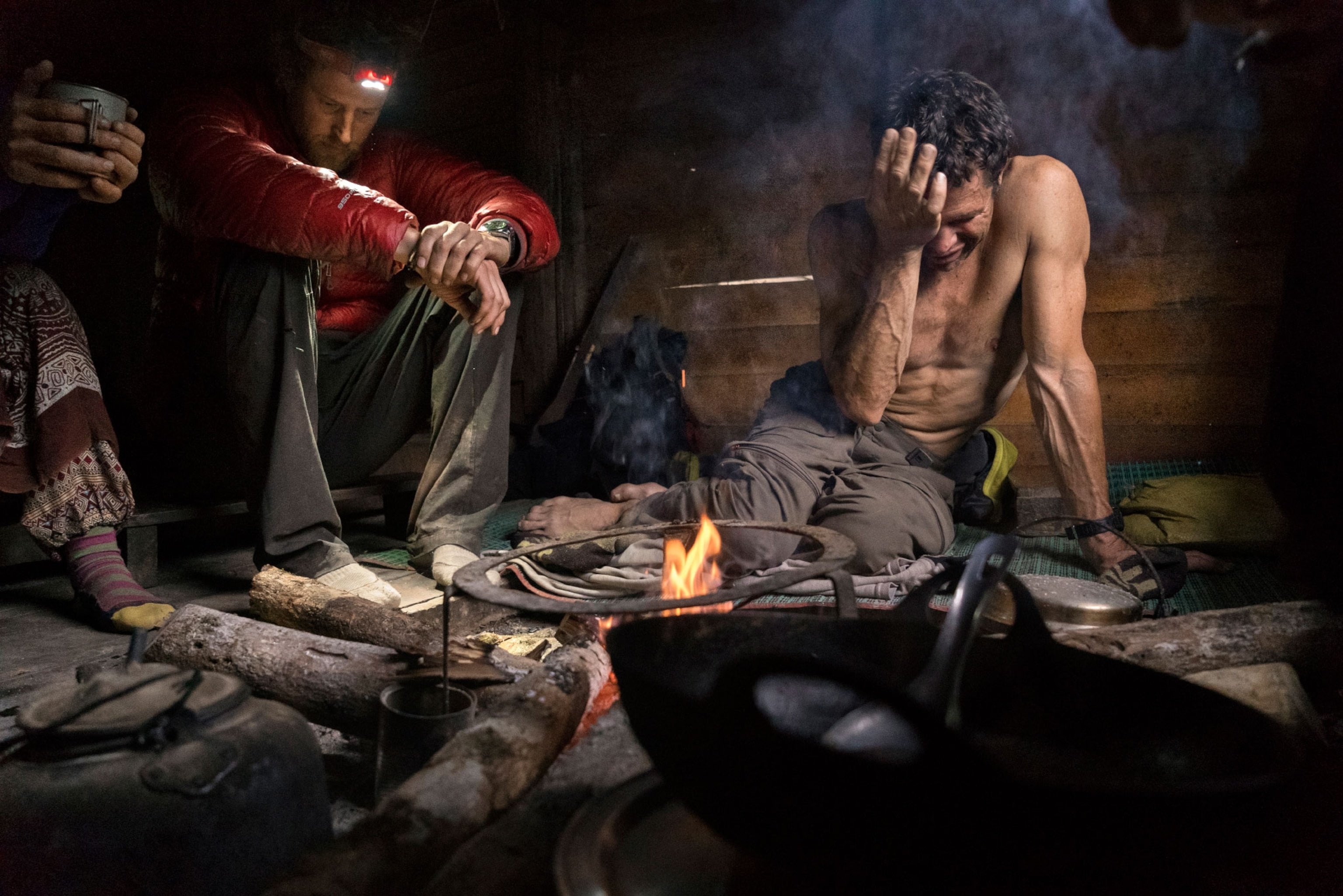three people sitting inside of a tent around a small fire, one of the men has his head in his hands