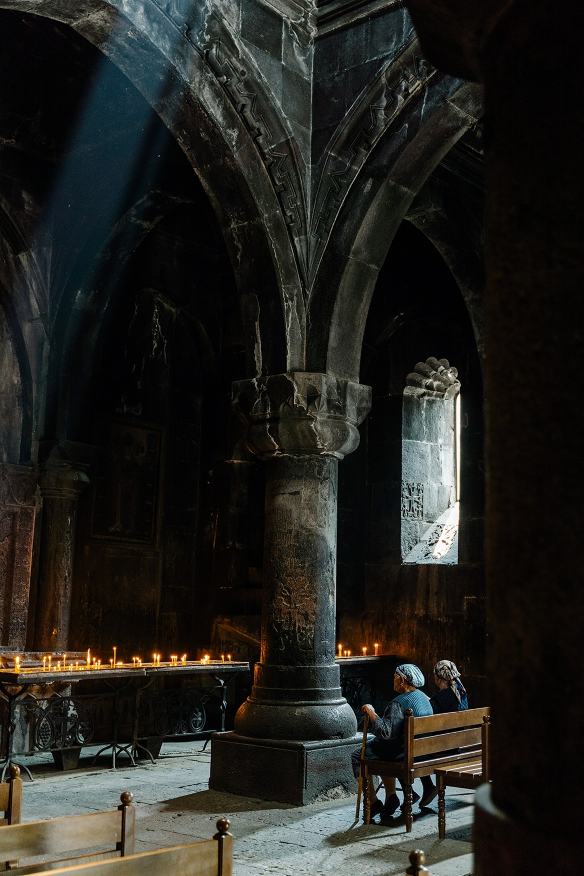 The interior of a medieval, stone church, with two elderly women sitting on a wooden bench in the front next to a towering pillar, looking at a candle-lit altar.