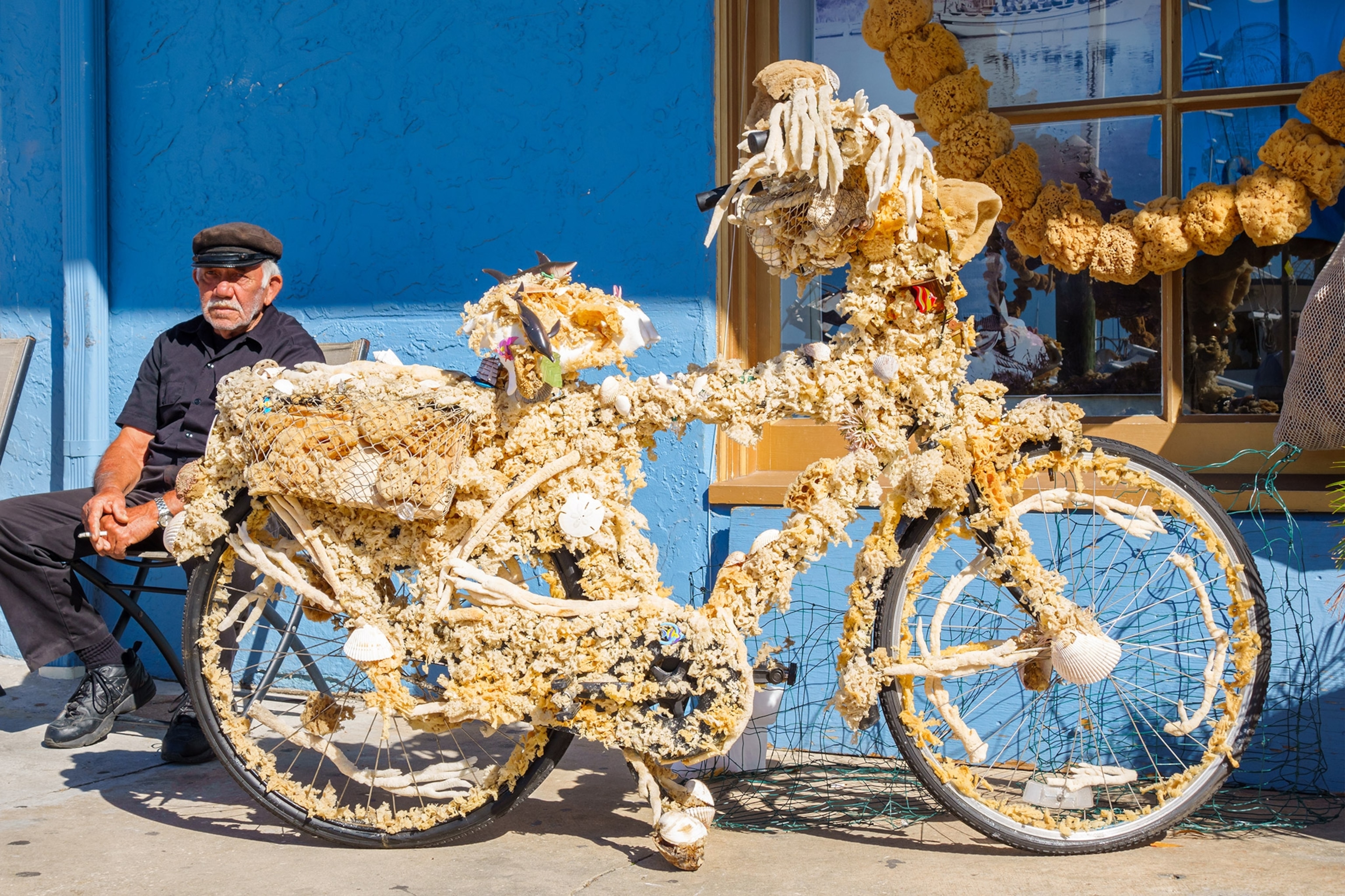 A senior man sits against a blue wall, a bike laden with dried sponges next to him