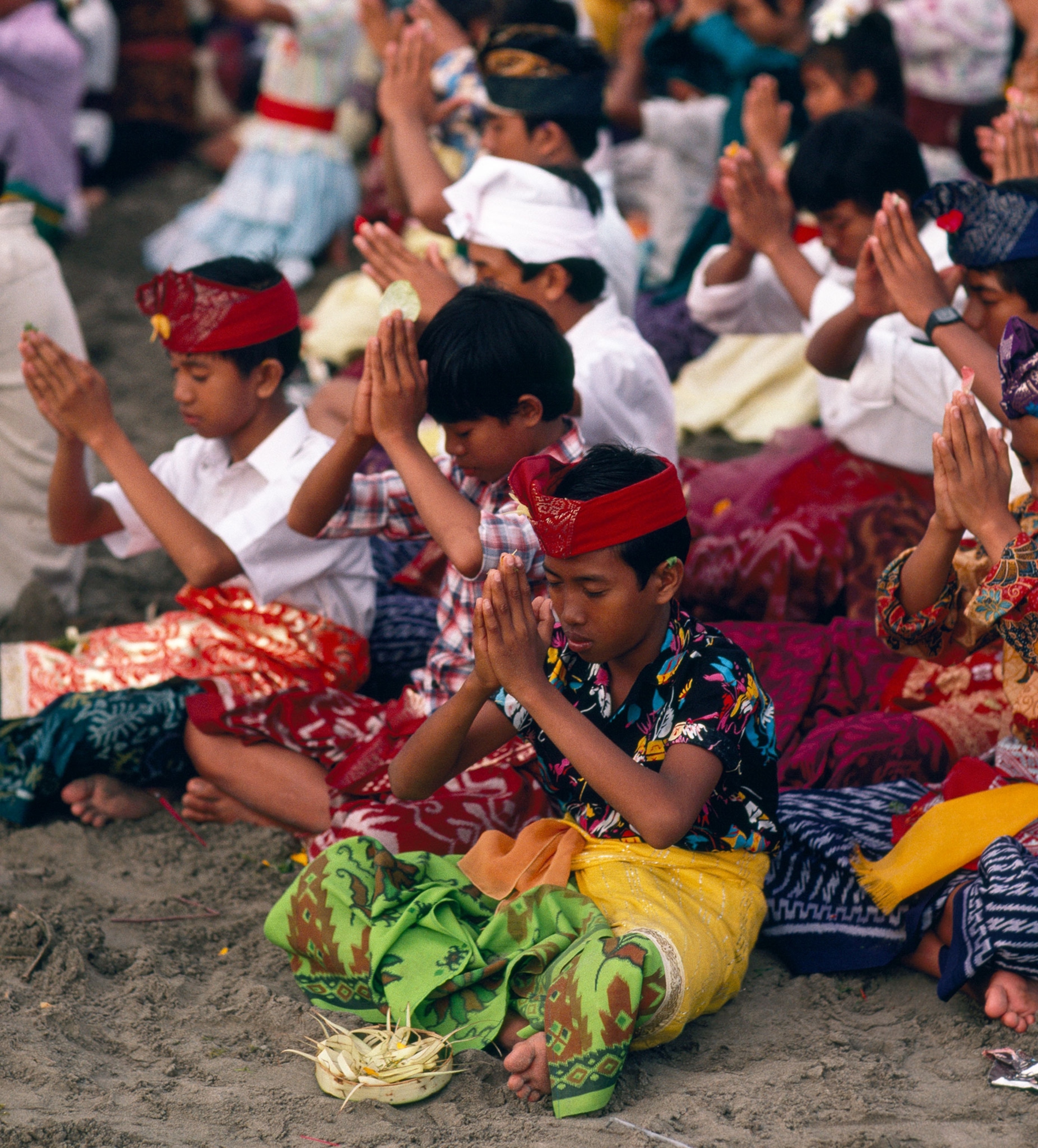 A group of young men dressed in colorful fabrics, sit crossed legged and pray on the beach.