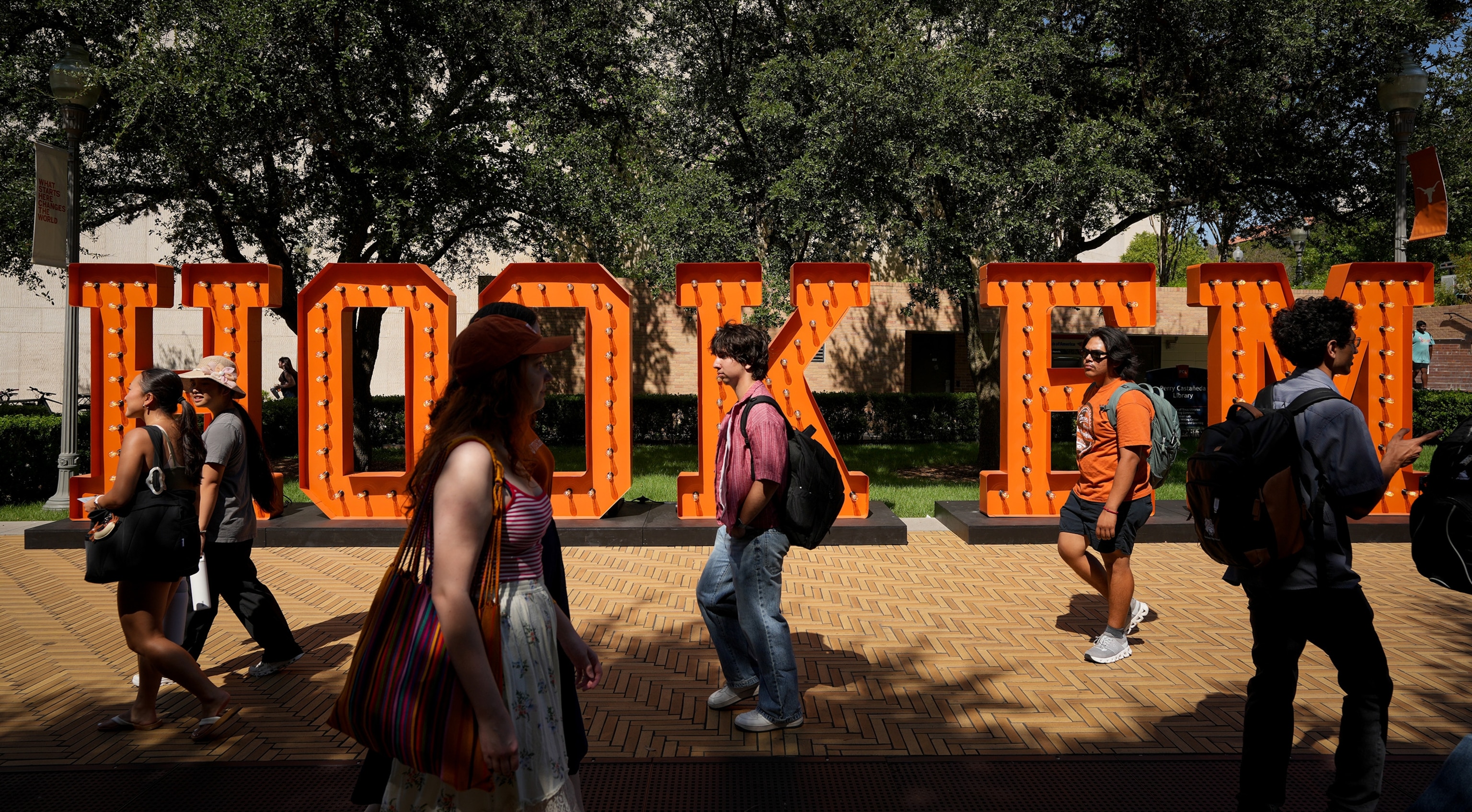 Students wearing backpacks and holding totes walk past large orange letters spelling out "Hook em"