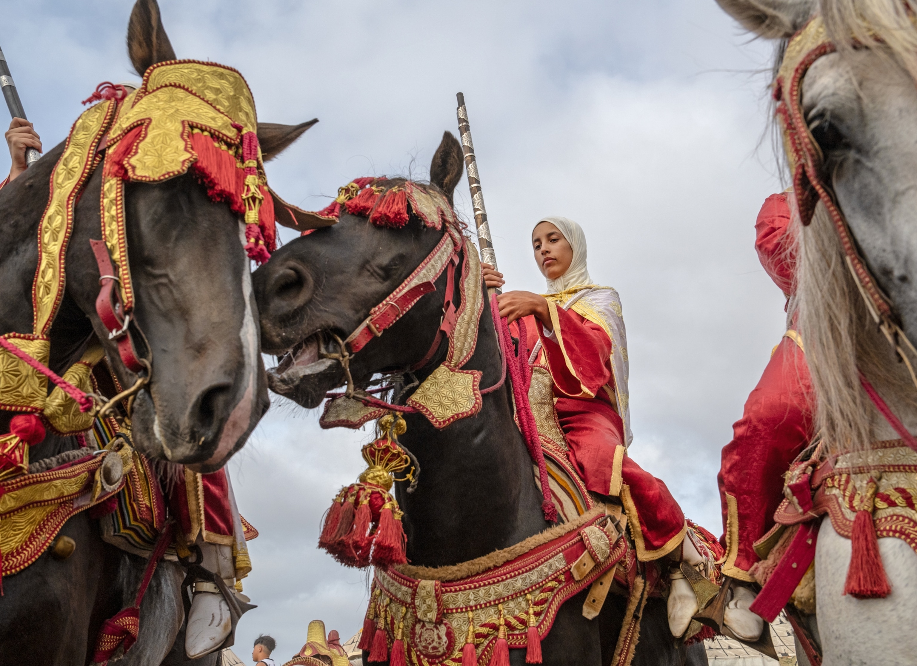 A look up into horses wearing gold and red tacks that go around their heads and chest. Women holding their guns and wearing red and white sit on their backs