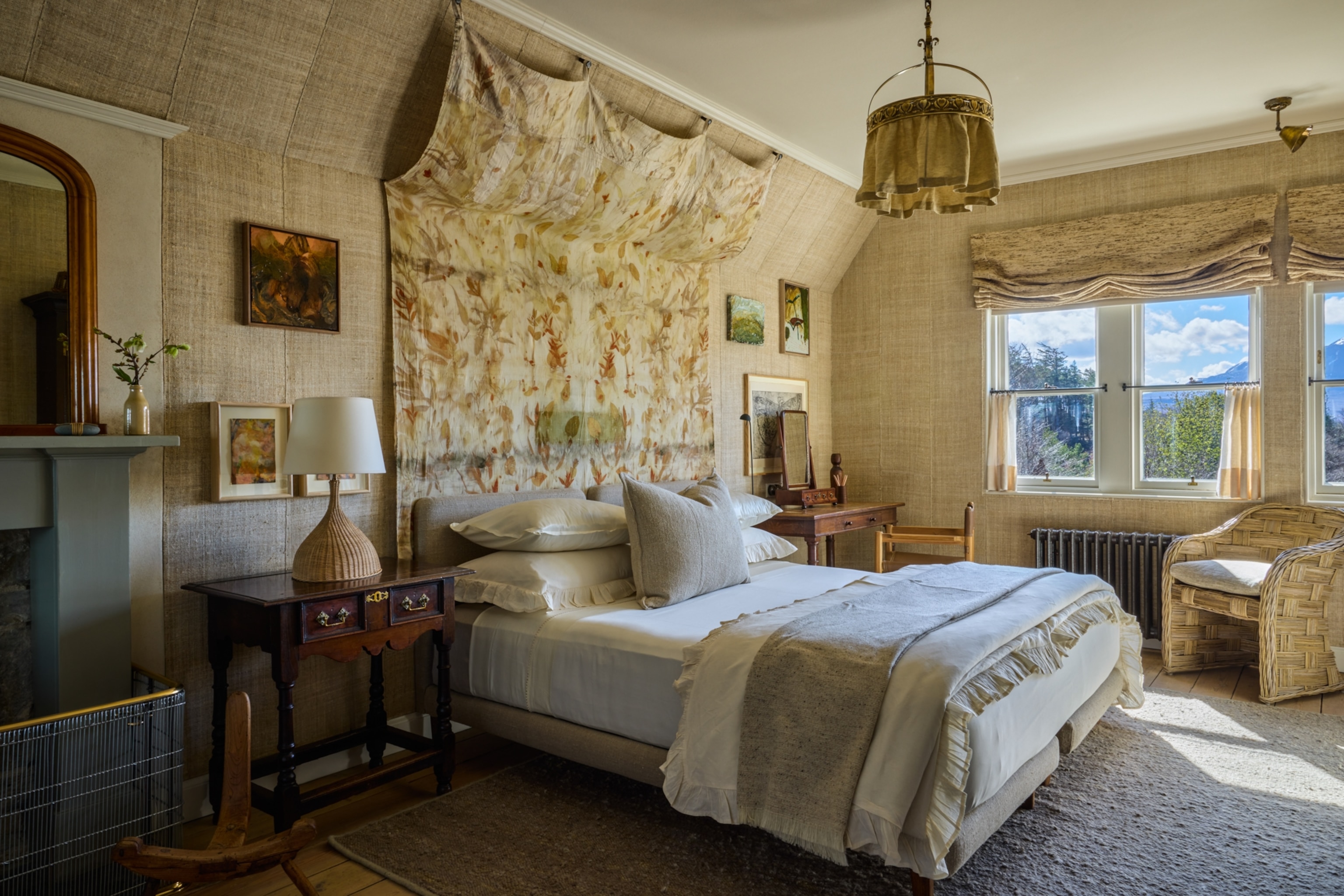 A cozy bedroom with a neatly made bed, framed artwork, wooden furniture, and windows overlooking a scenic outdoor view.