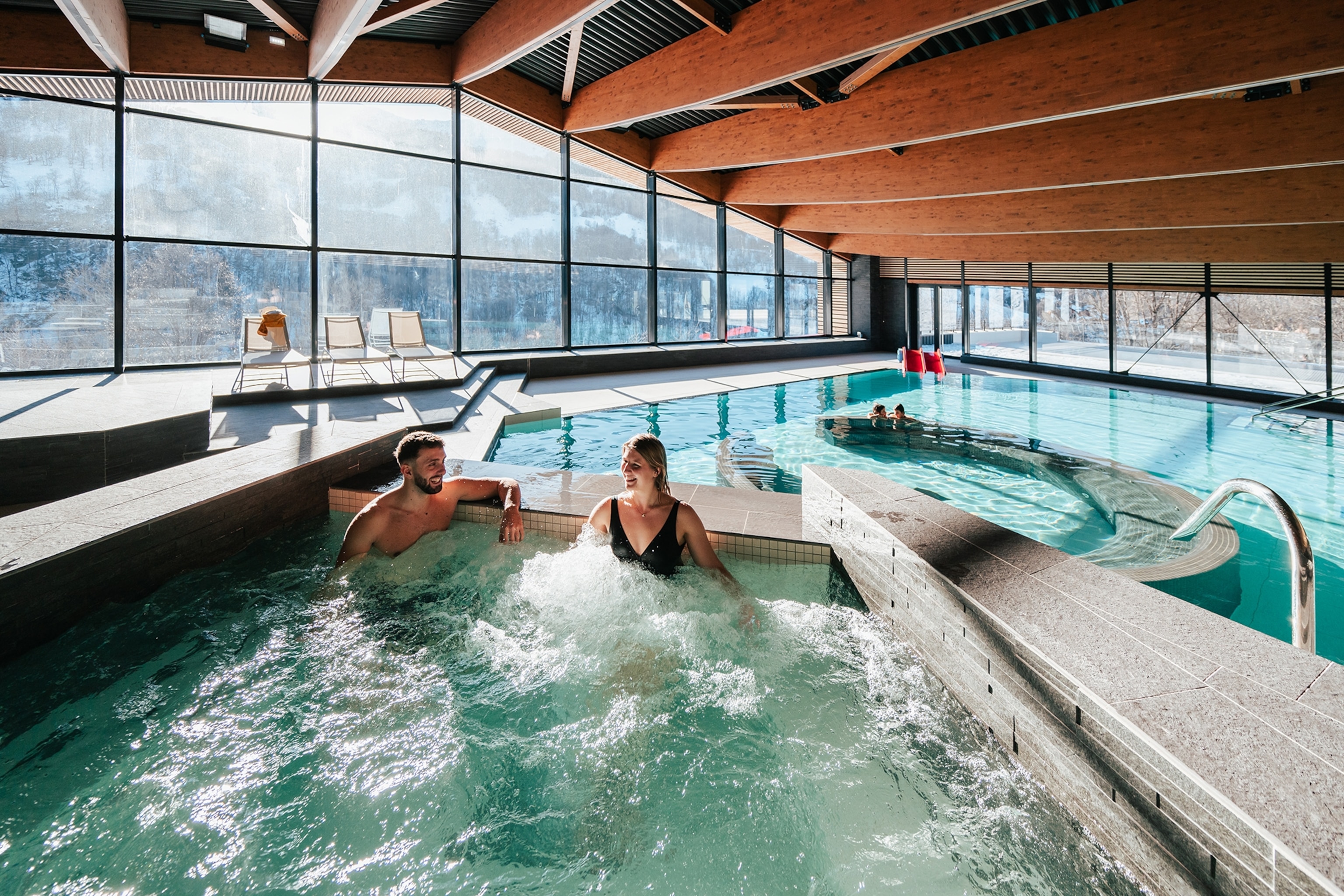 Two visitors of a spa and wellness centre with a glass front to the Alps relaxing in a jacuzzi.