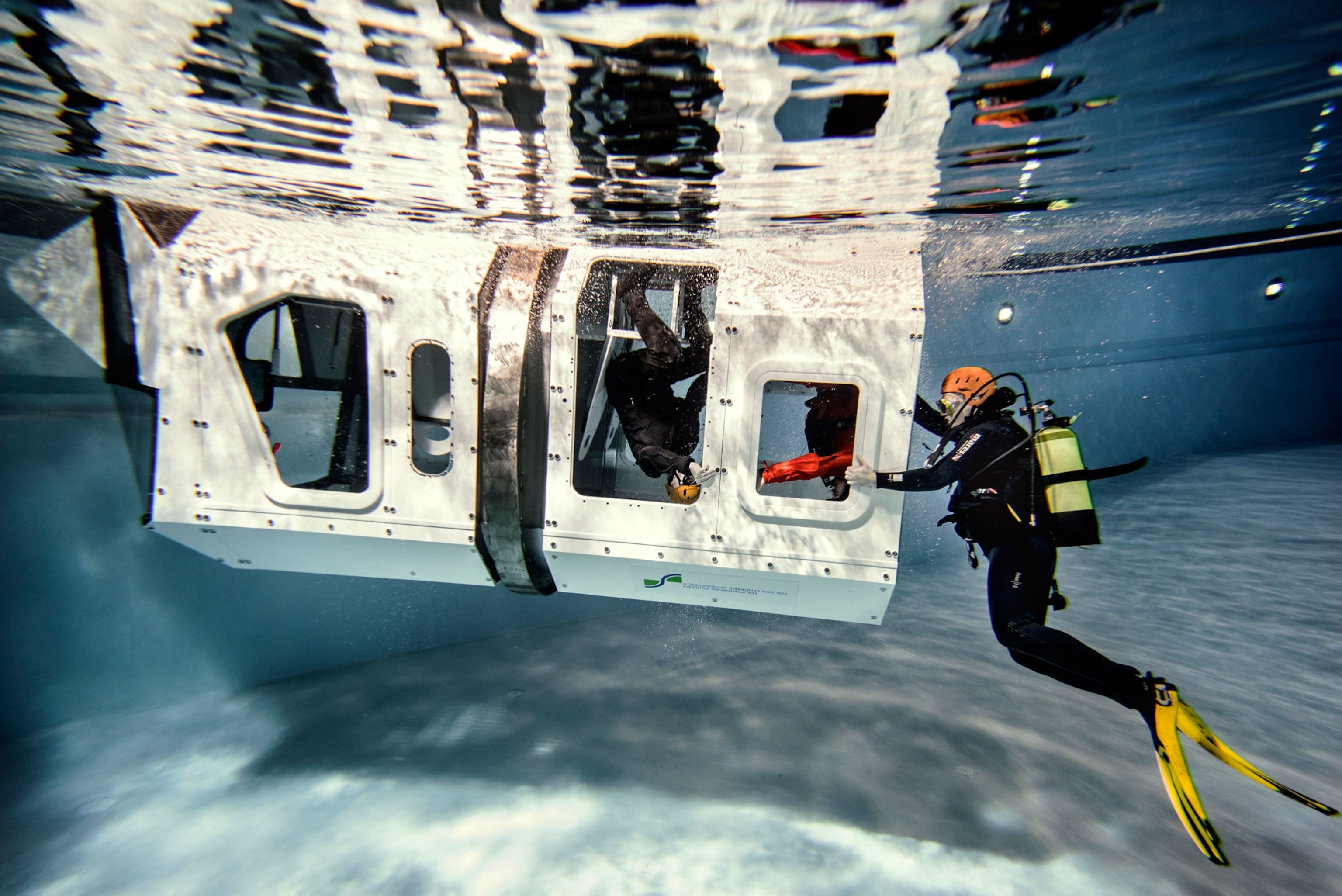Underwater picture of an instructor and a safety diver watching an upside down trainee try to escape from a capsized-helicopter simulator