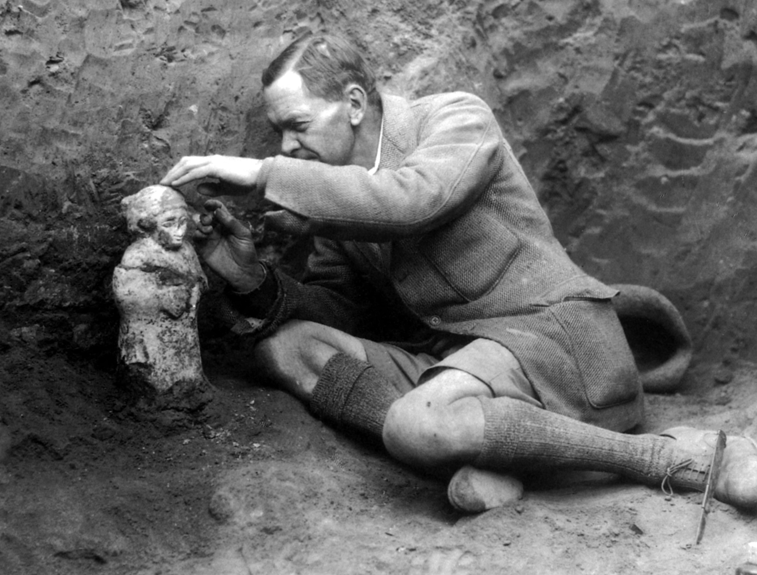 A man in an archaeological dig sits on the ground, carefully dusting a small ancient statue. He wears a tweed jacket and shorts