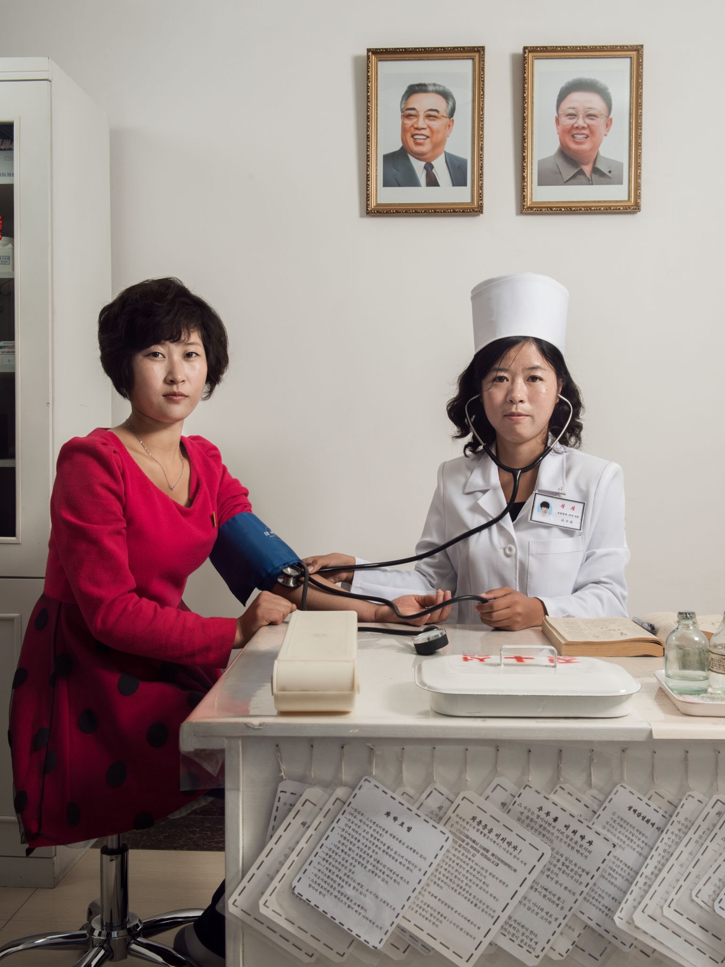 two women, a doctor and a worker posing for a portrait, two frames hang on the wall