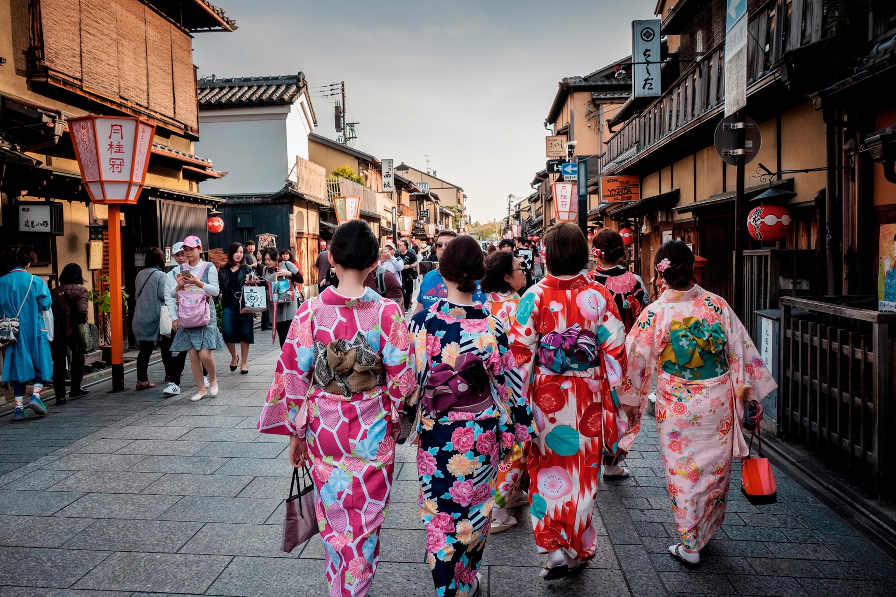 Four traditionally-dressed geishas, seen from behind, walking down a busy street in the ancient town of Kyoto.
