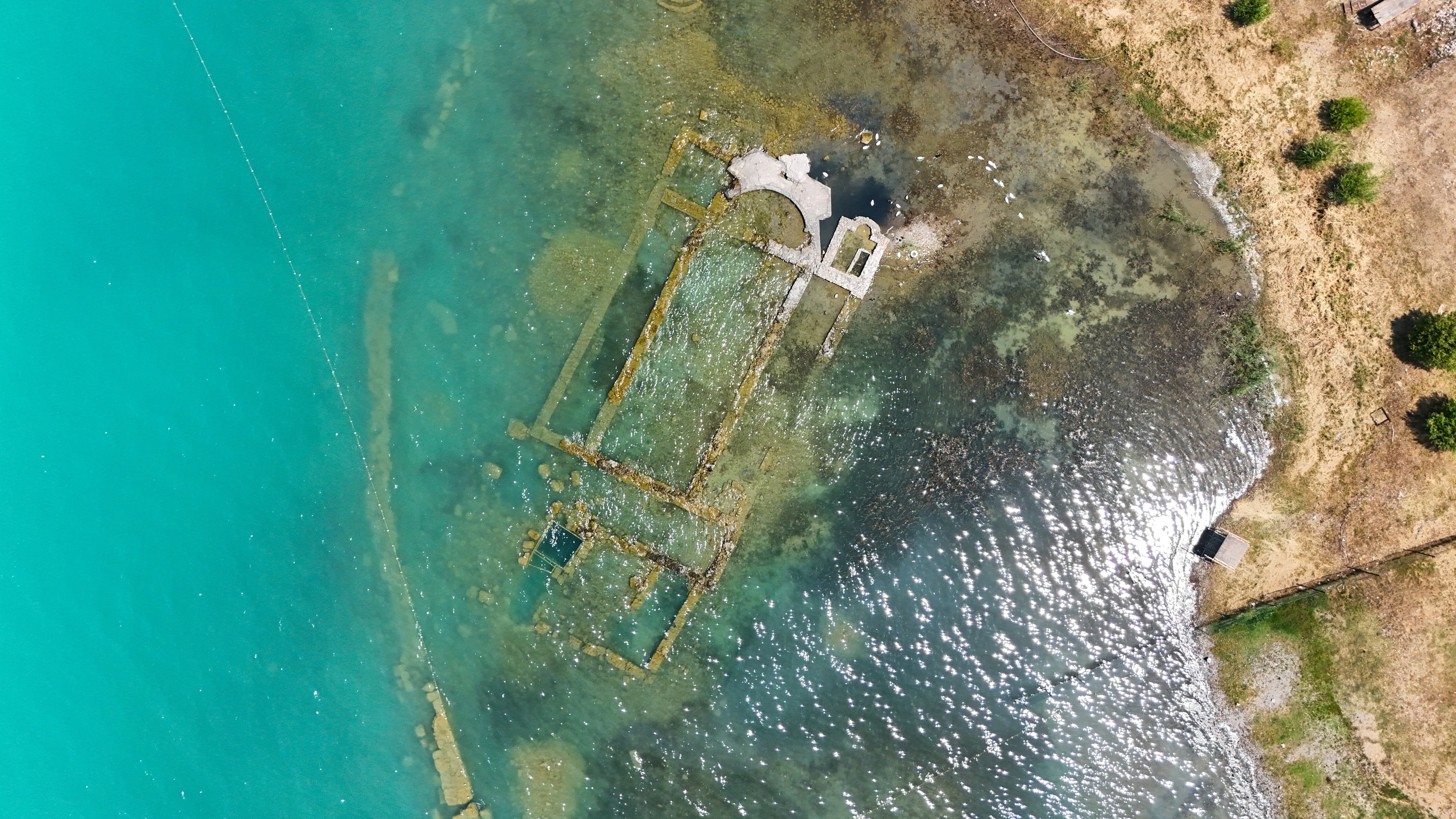 Arial photo over a blue lake with a cement structure emerging from the water near the shore.