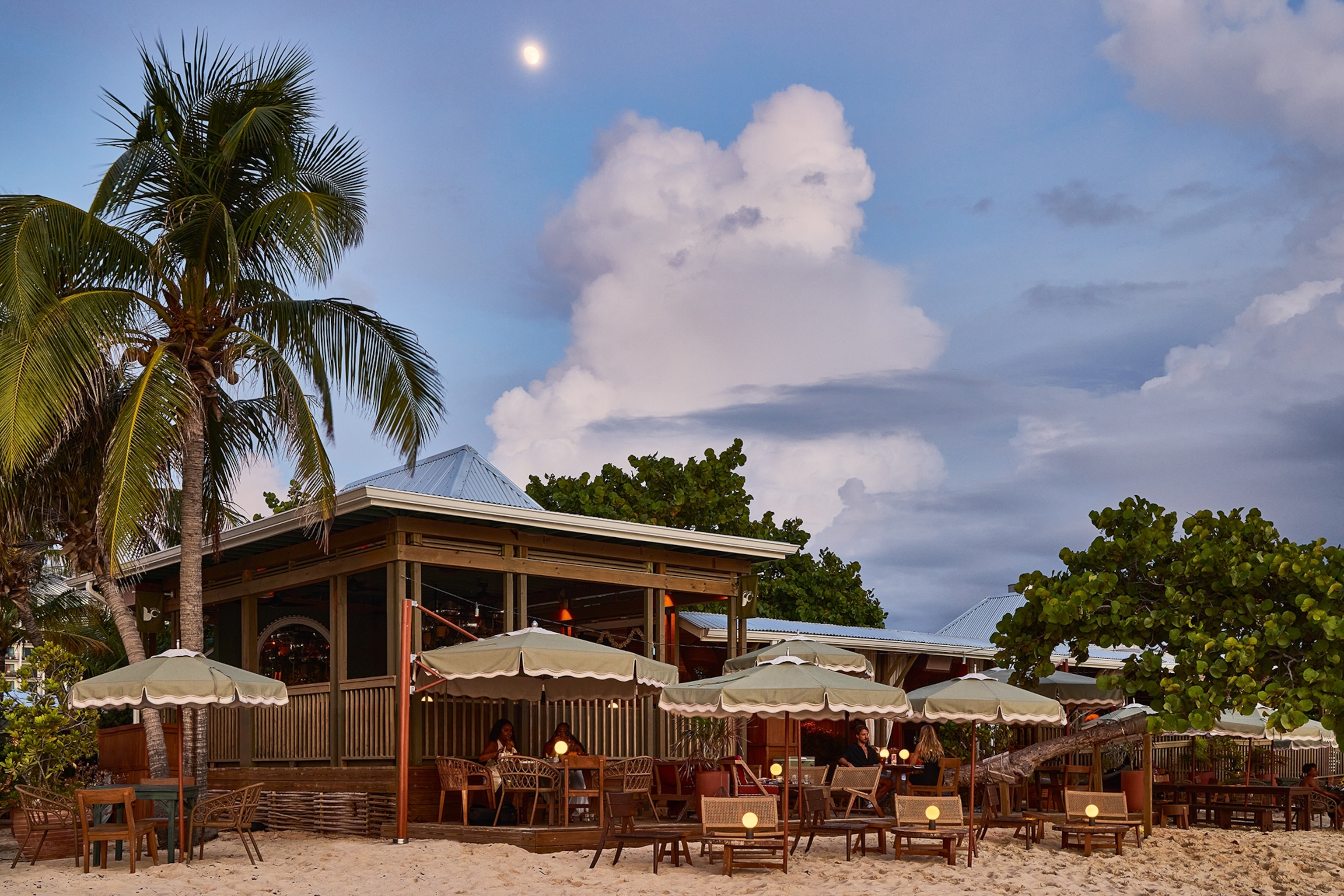 A beach side hut and terrace with sunchairs and parasols, surrounded by palm trees.