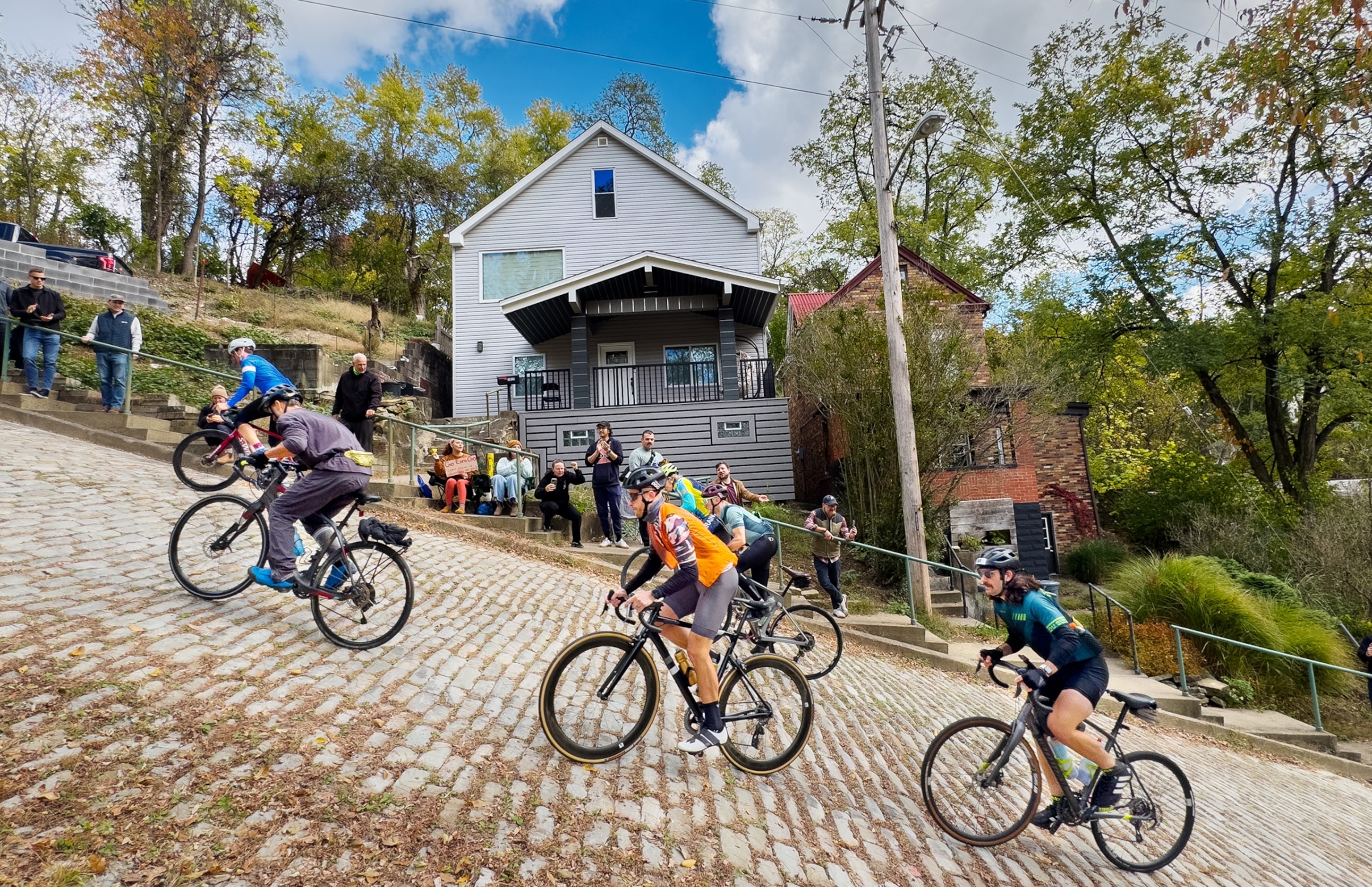 Cyclists lean off their seats as they push their bicycles up a steep hill. In the background is a house and people lining the sidewalks to watch the cyclists make their way up.