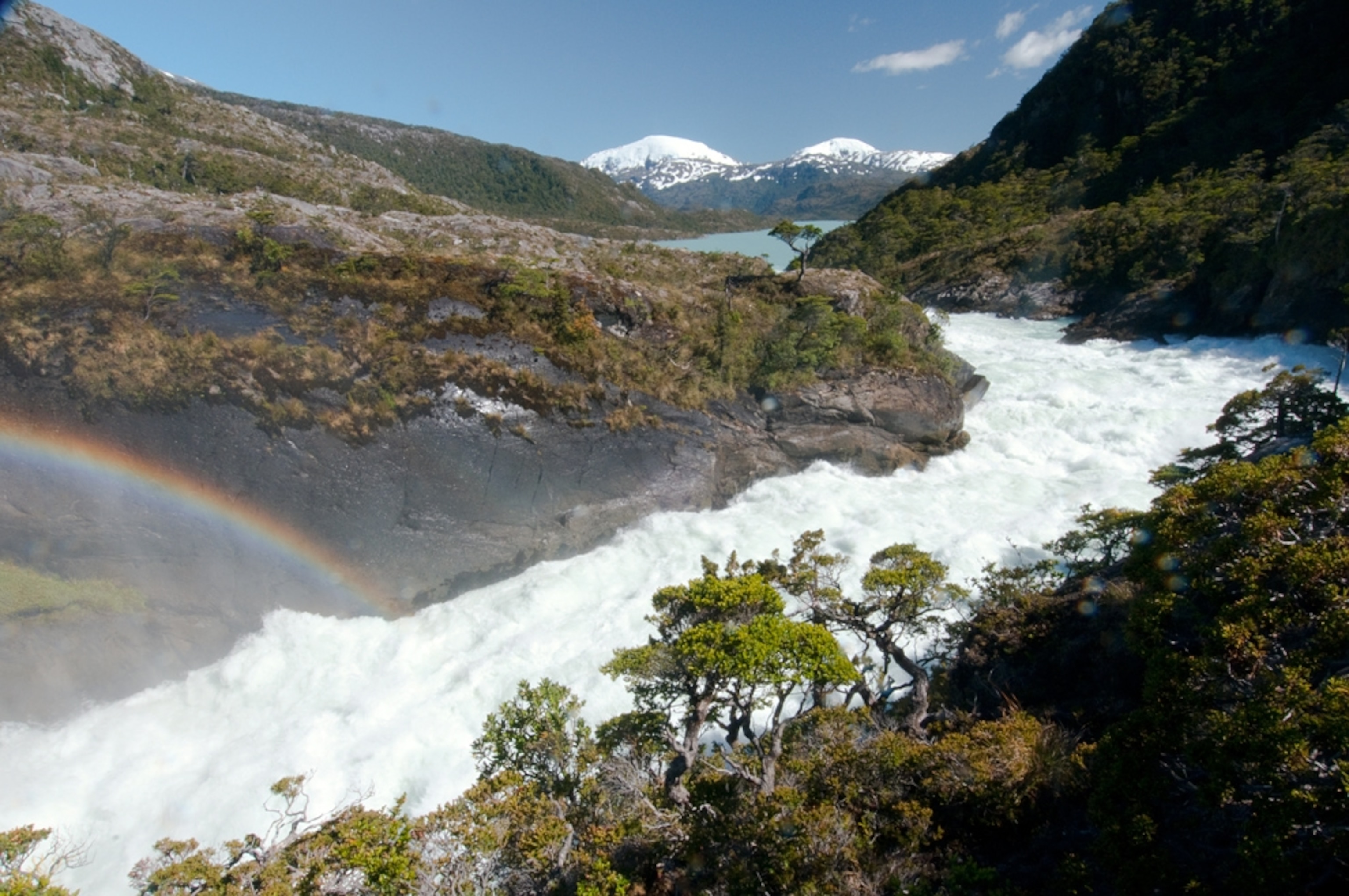 A rainbow appears above river rapids, fed by snowcapped peaks in the background.