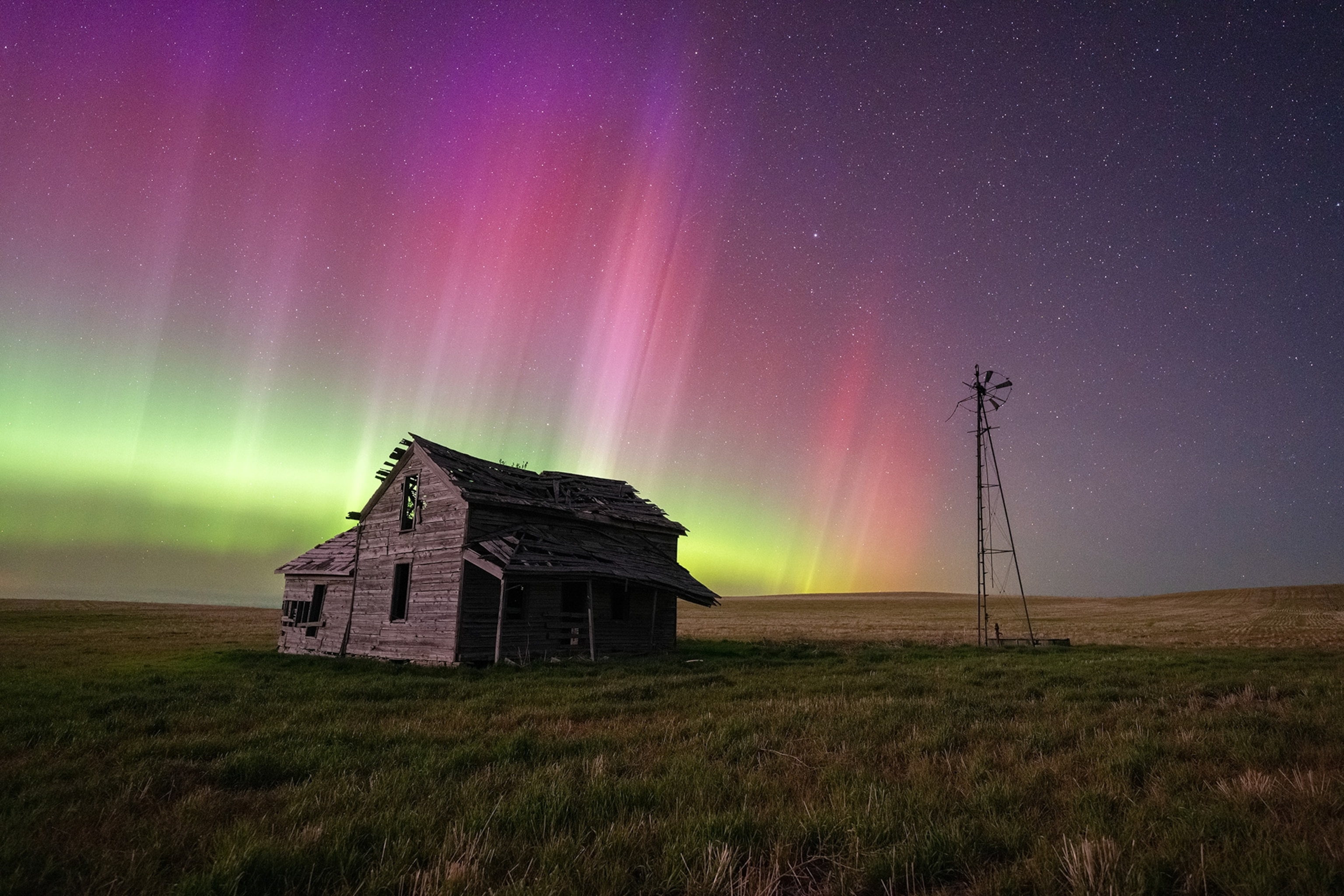 A green, pink, and purple evening sky sits behind an old wooden house in the middle of a field.