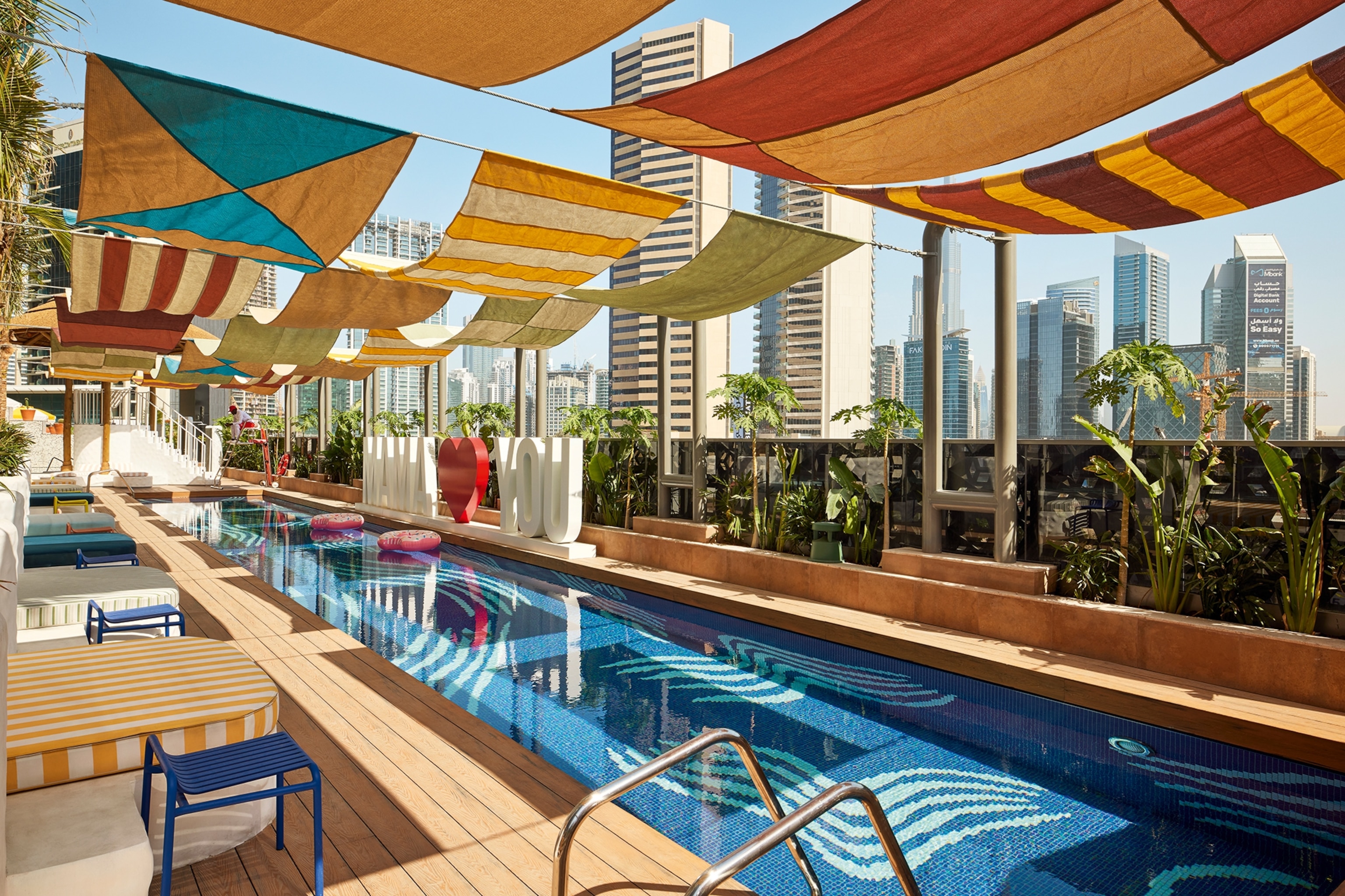 A rooftop pool with funky mosaic and square panels of fabric tied together above to lend shade.