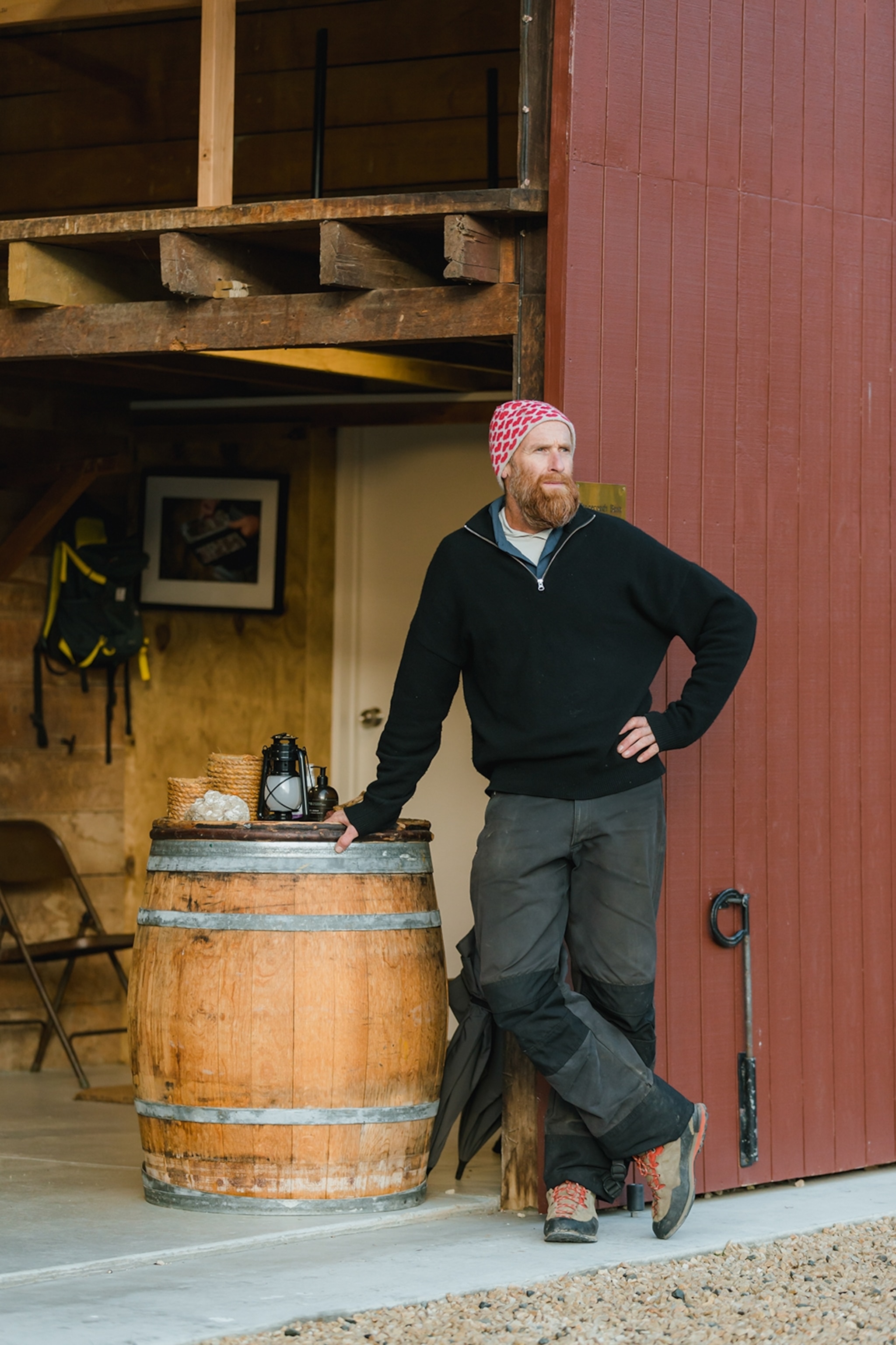 A bearded local casually leaning against a barrel at the entrance to a barn,looking into the distance.