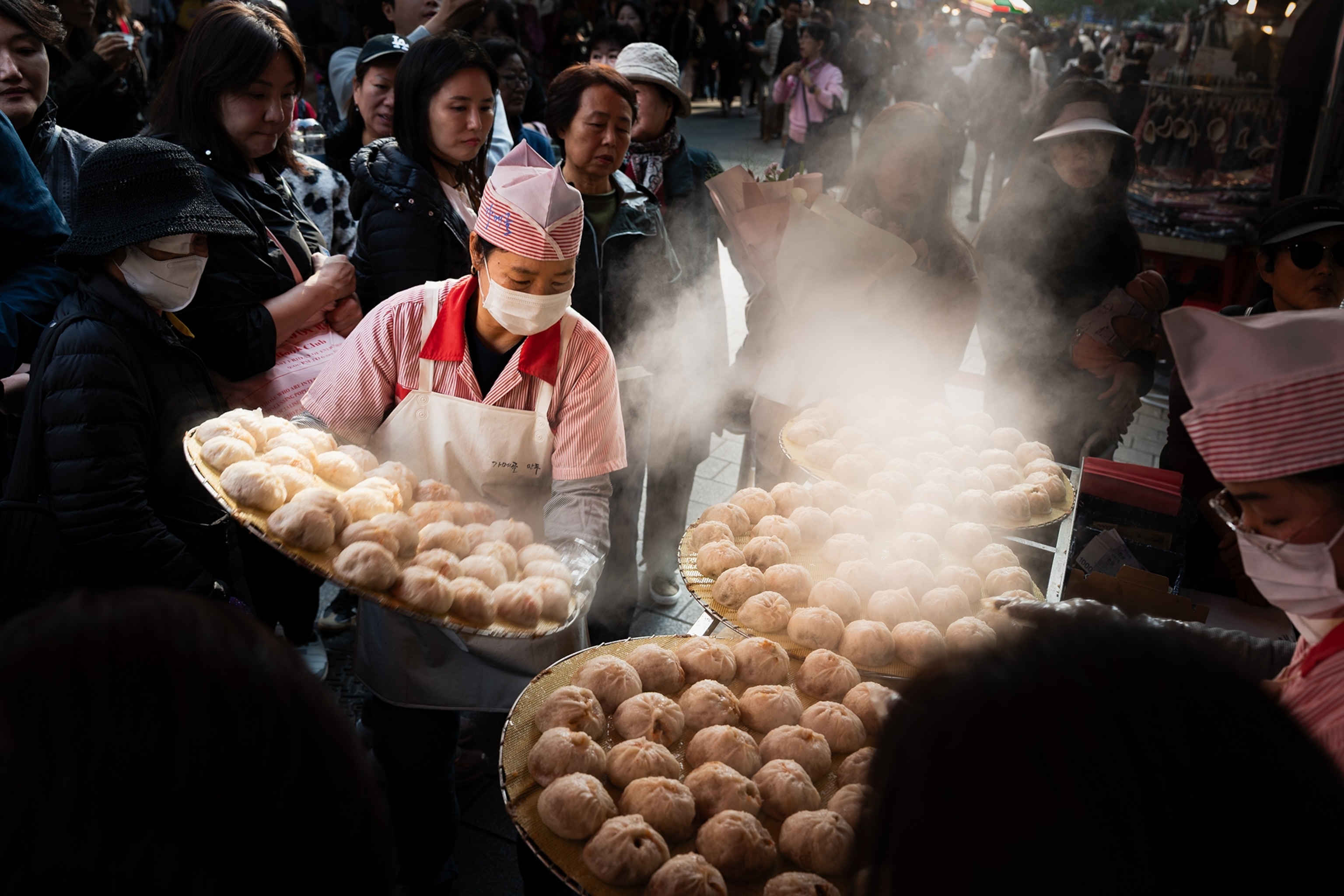 A steamed bun seller on a busy market in Seoul carrying a full tray past waiting customers.