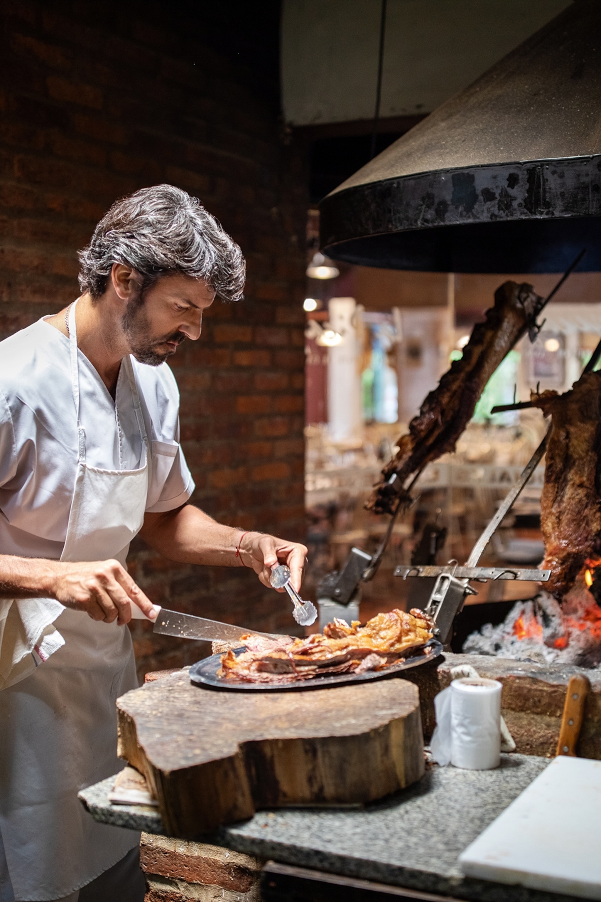 A young chef cutting a wood-grilled steak on a silver platter next to gleaming embers and meat on a spit.