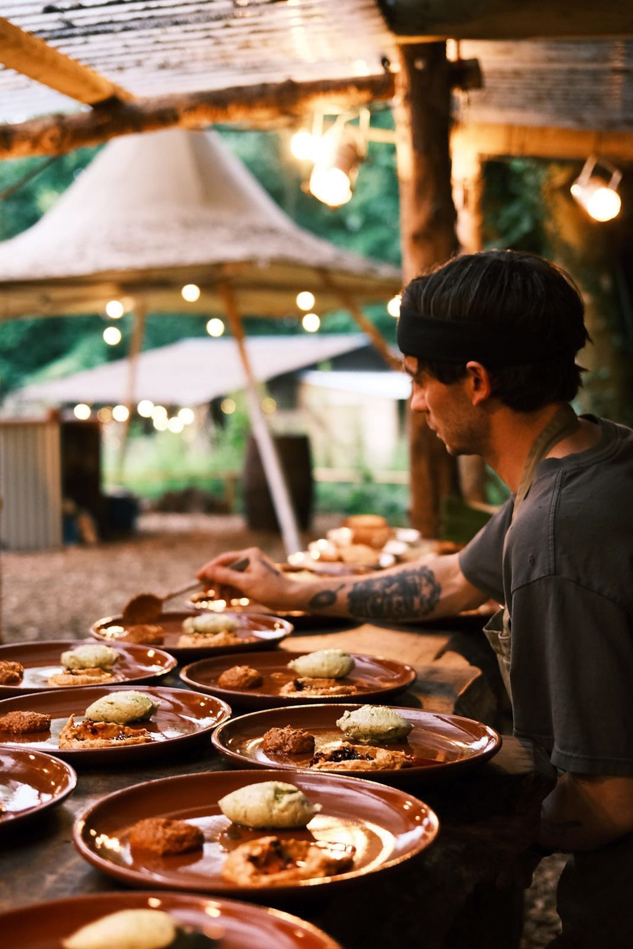 A young chef plating up food on an outdoor pass in a campsite illuminated by fairy lights.