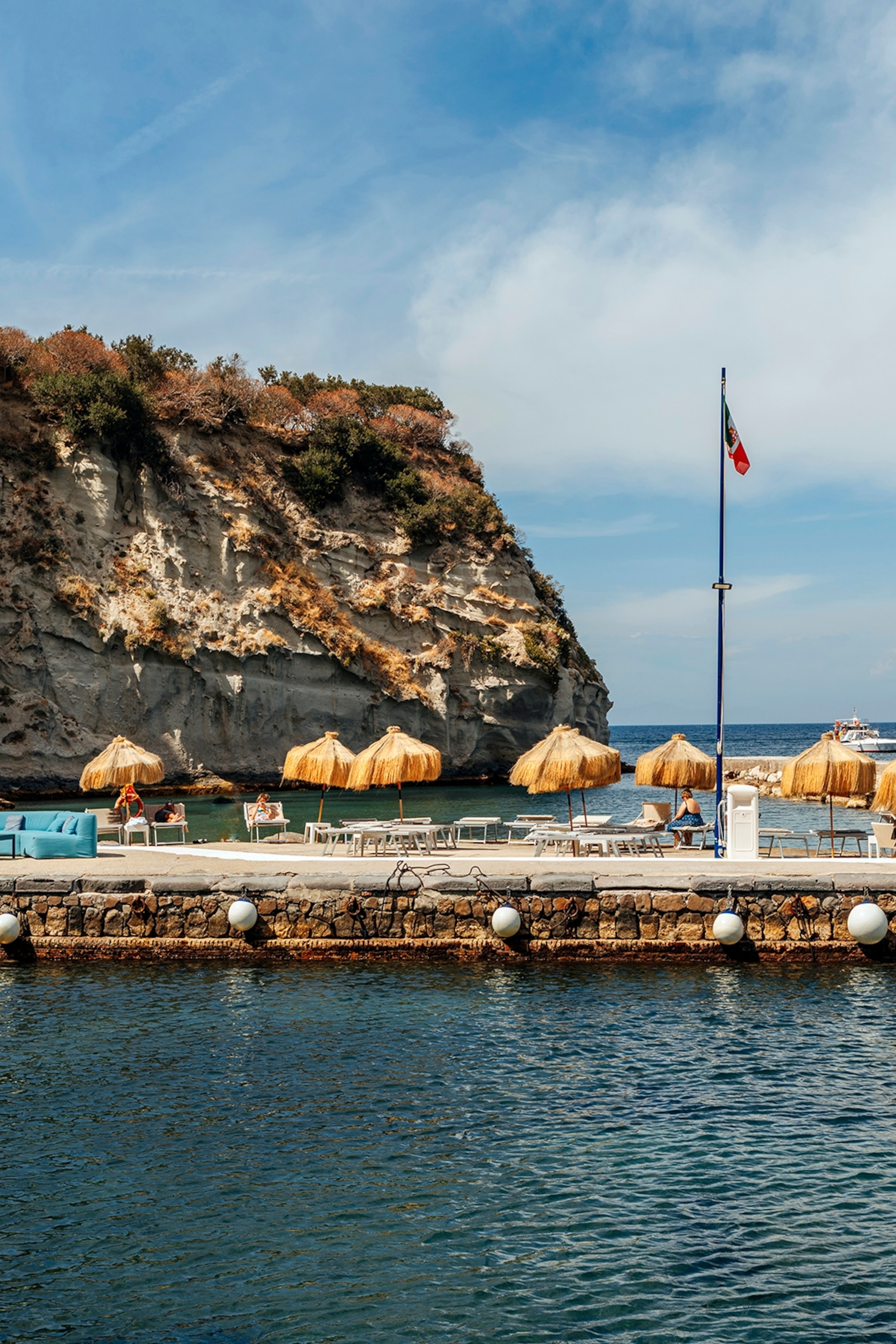 A concrete boardwalk with sun chairs and parasols as bathers jump into the sea below.