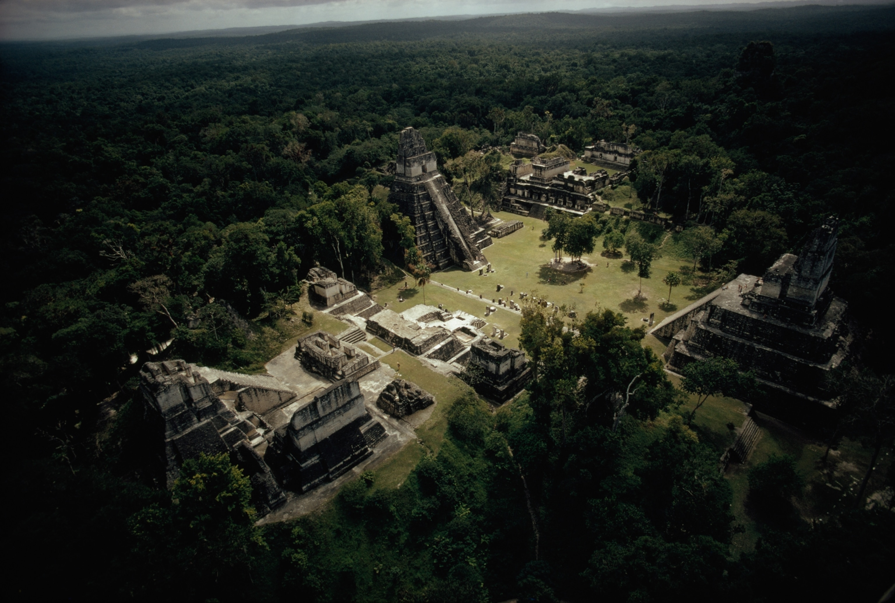 Aerial view of ancient Mayan ruins surrounded by dense green forest, featuring large stepped pyramids and scattered stone structures under a cloudy sky.