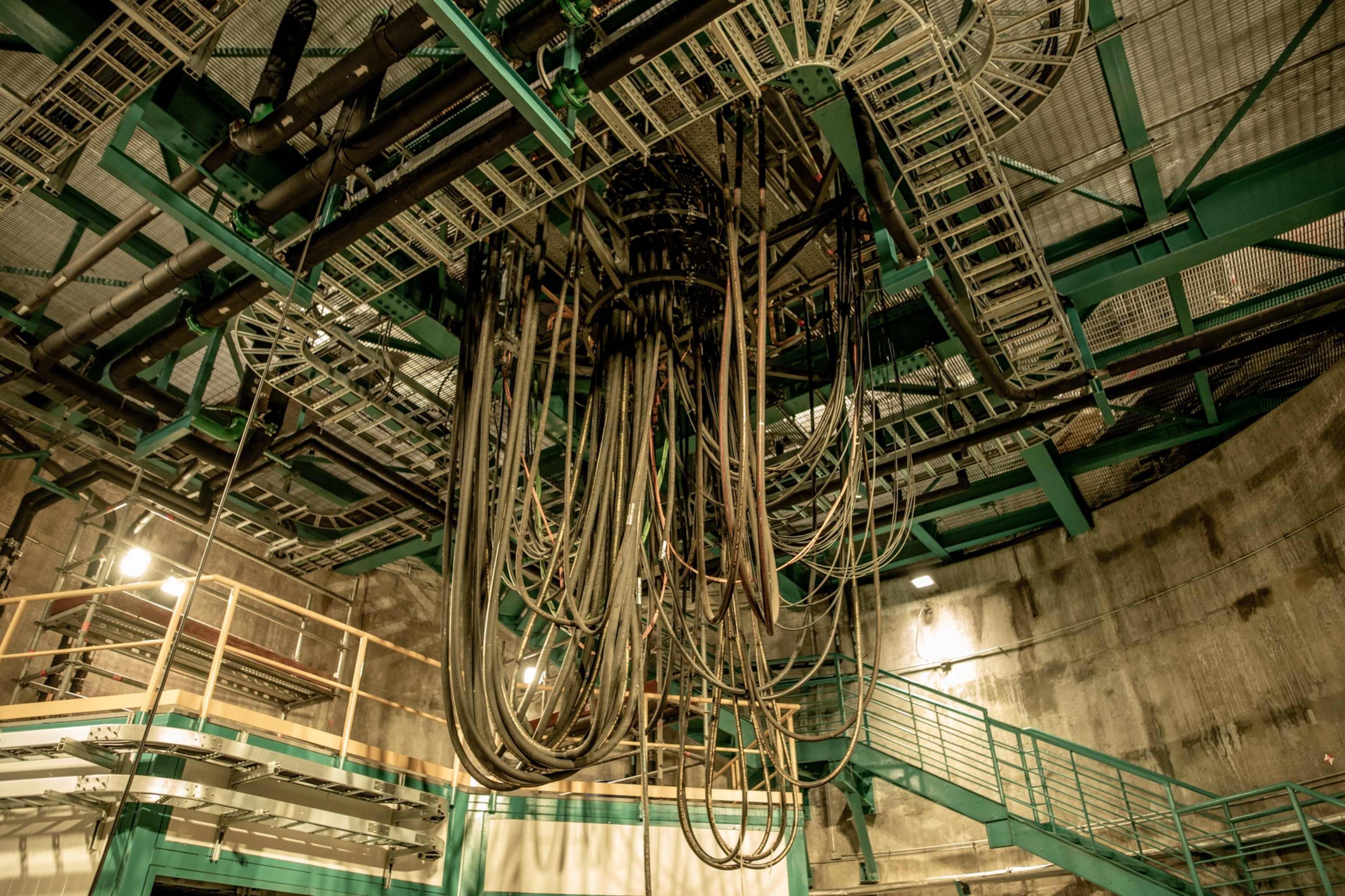 An enormous tangle of thick cables hanging from an industrial ceiling surrounded by catwalks