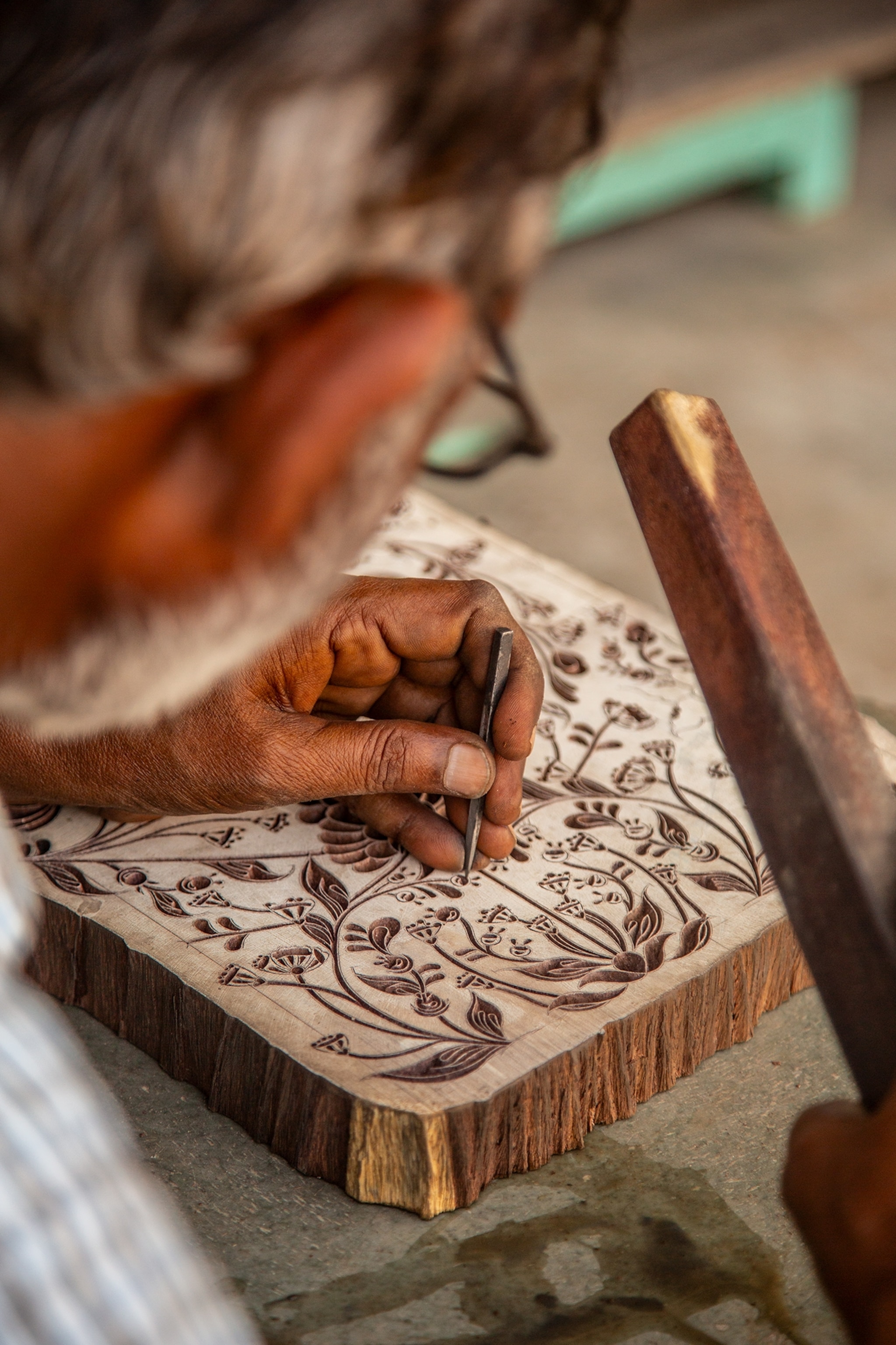 A craftsperson uses a small carving tool to create an intricate floral design on a wooden block, with detailed patterns etched into the surface.