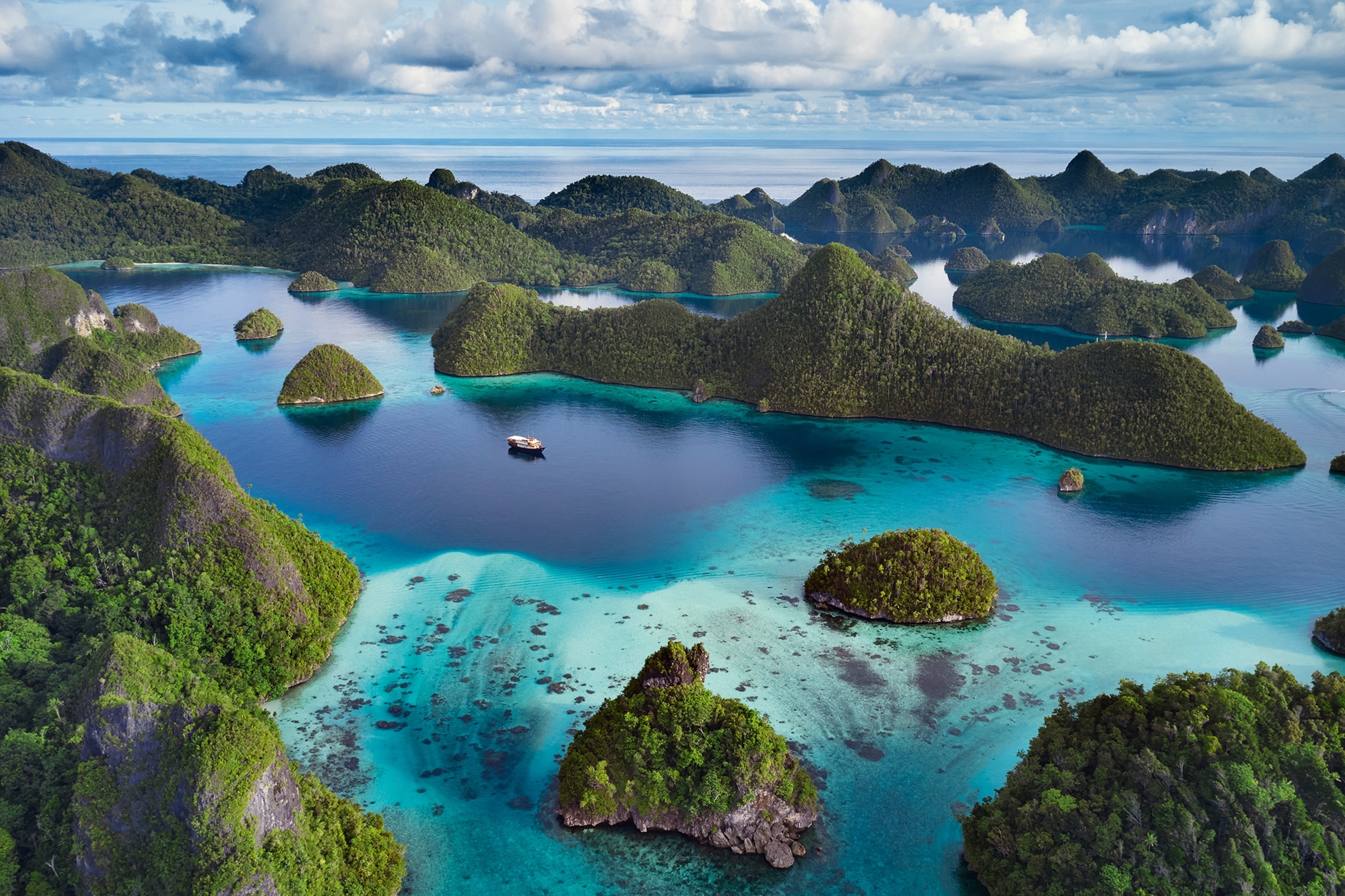 An aerial shot of mushroom-like, lush islands strewn across and ocean inlet with a sole boat in the centre.