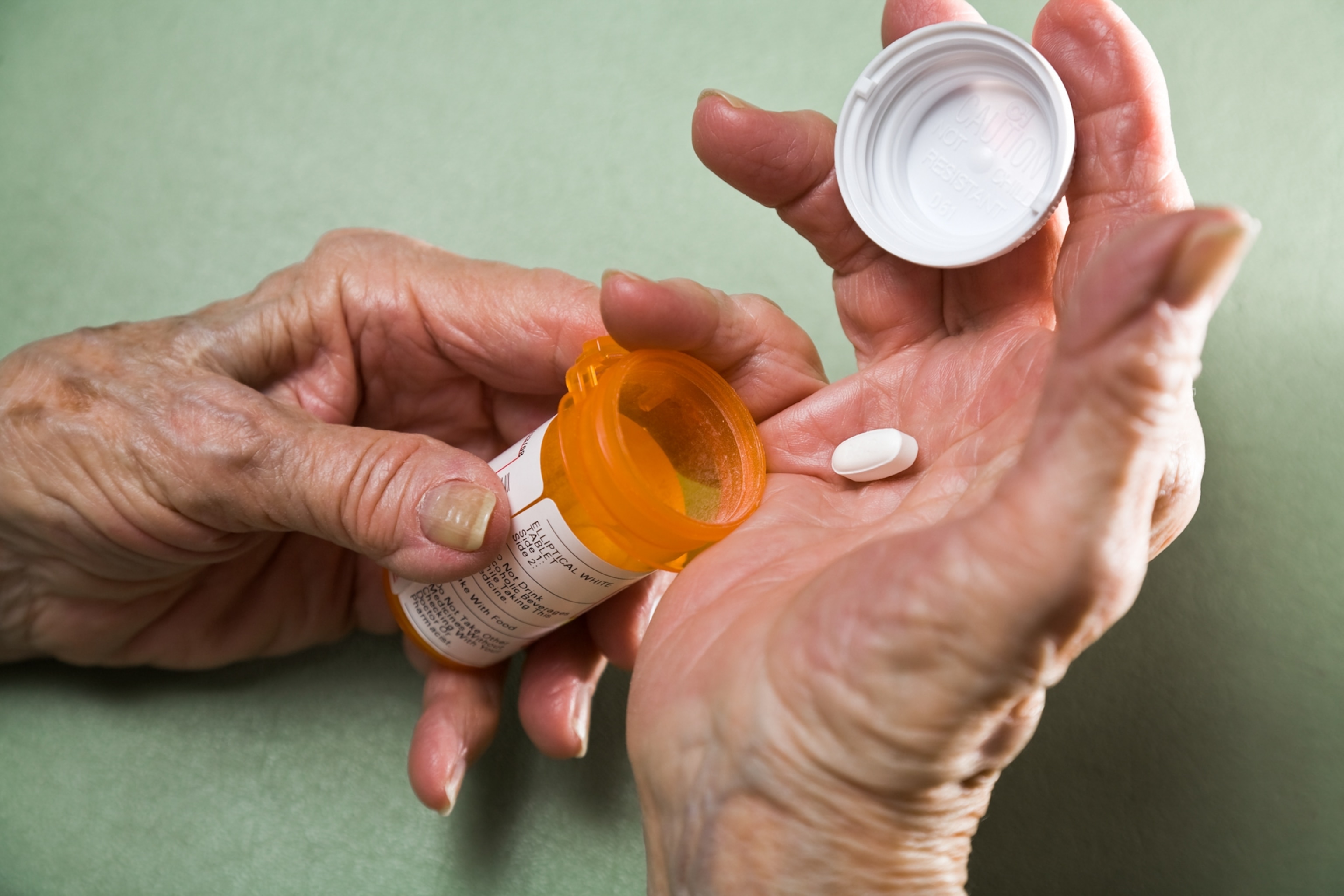 Elderly hands holding an orange pill bottle and dispensing a white pill into the palm, with a green background