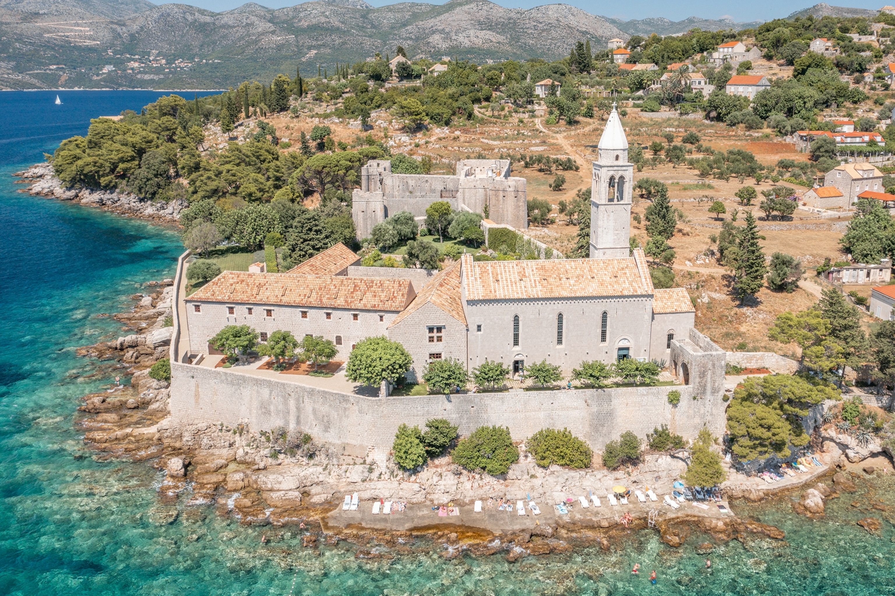 An aerial view onto an old stone Monastery on the edge of a rocky island, looking over the clear waters of the ocean.