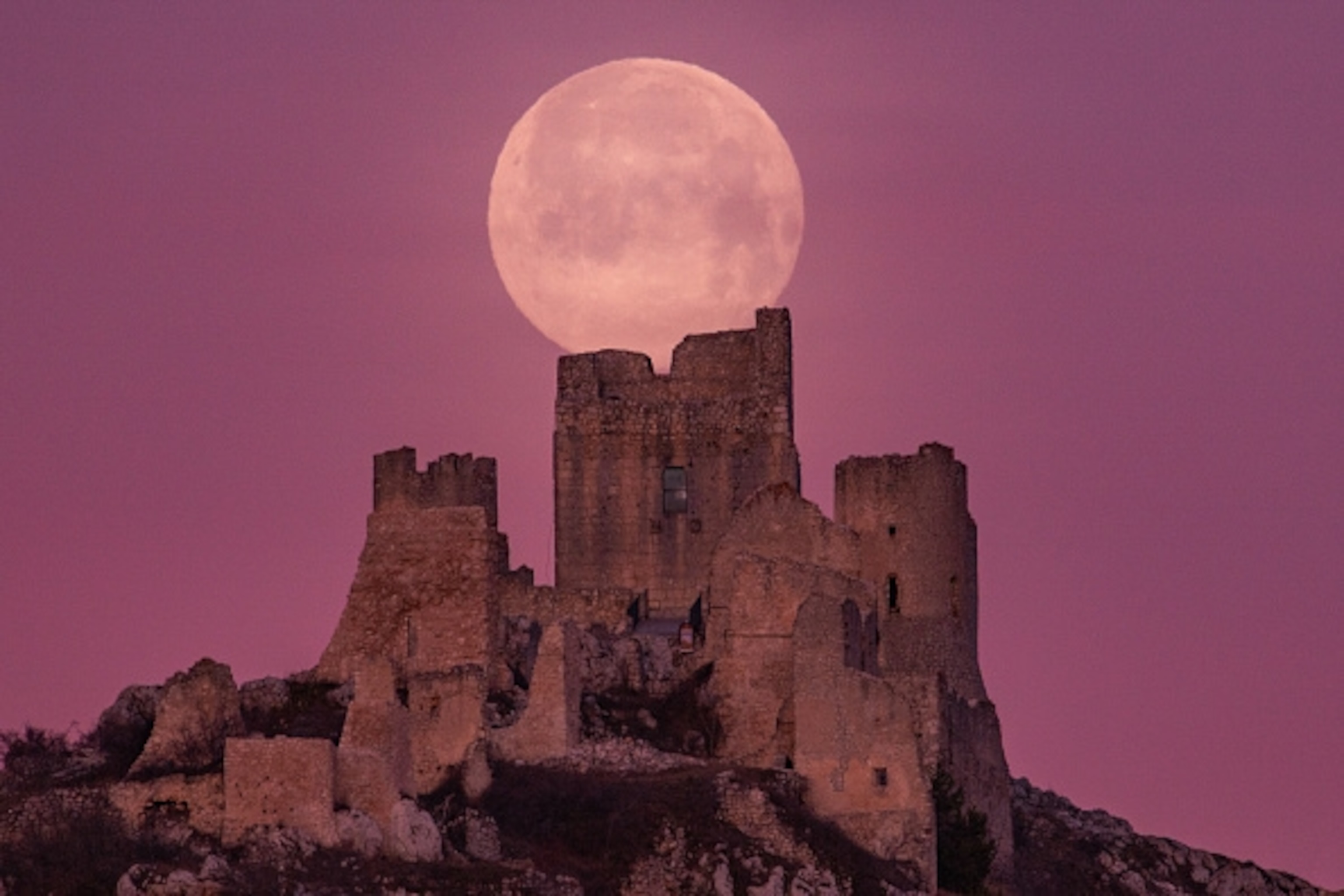 Ancient stone castle ruins silhouetted against a large, full moon and a vibrant pink-purple sky.