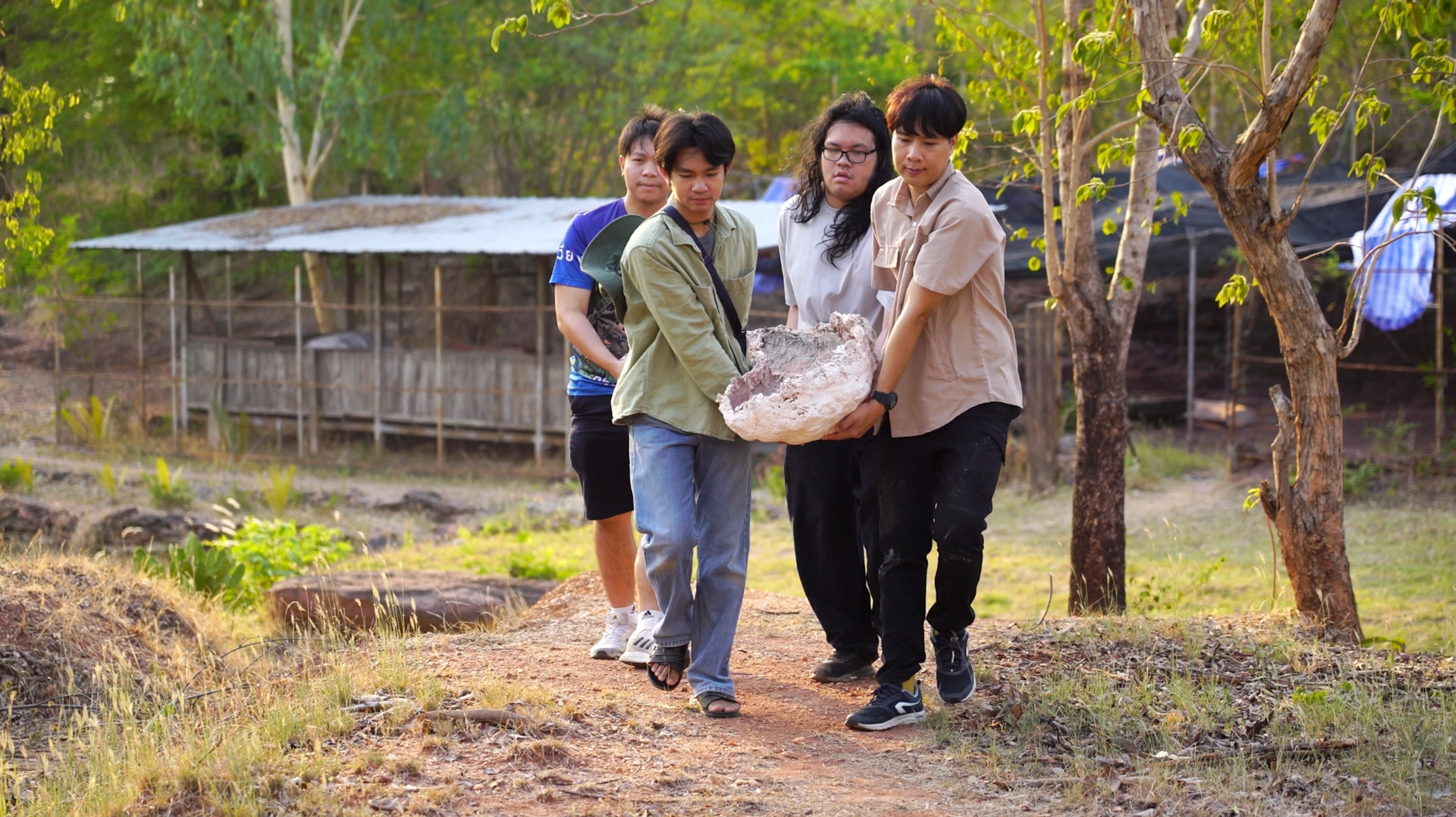 Four young men carrying a large fossil wrapped in cloth along a dirt path, flanked by trees and a rustic shelter